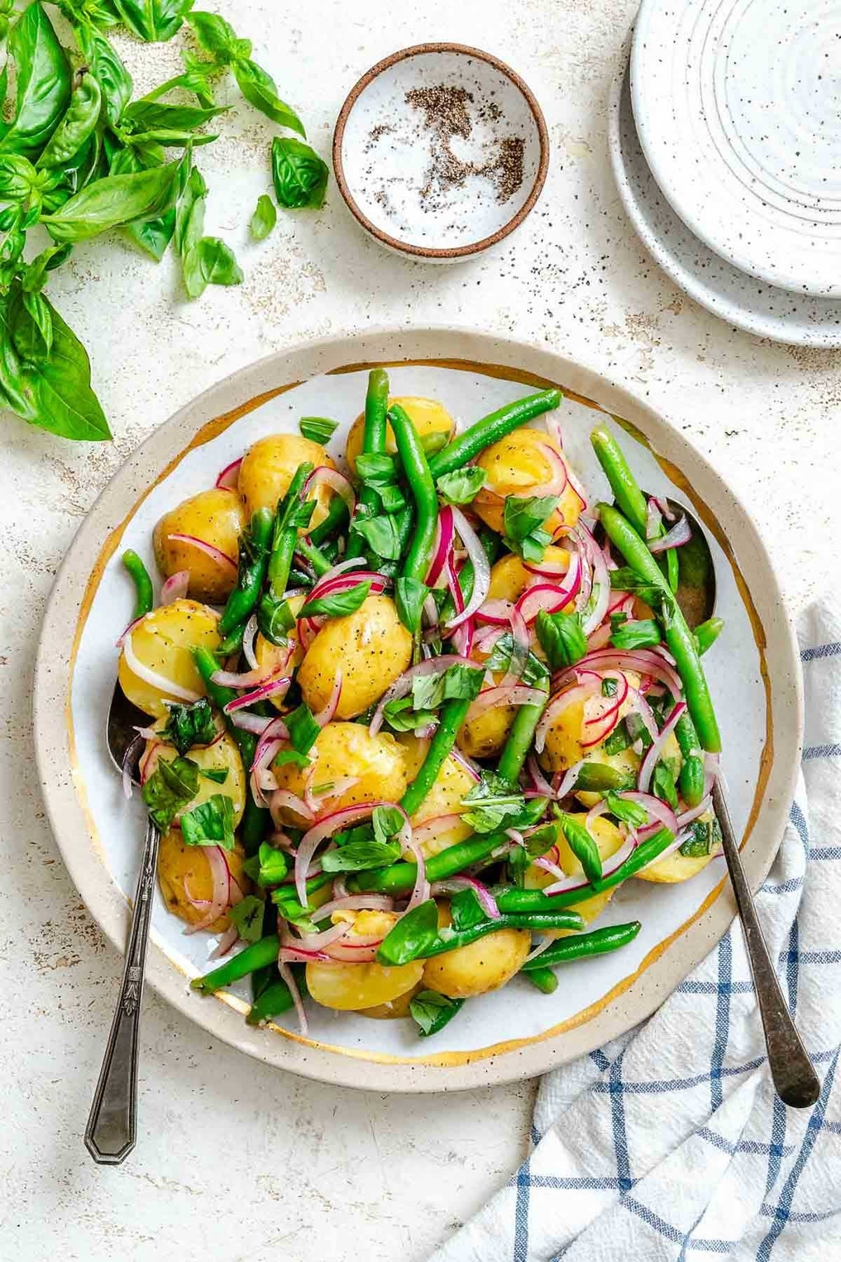 Potato salad with green beans, red onions, and fresh herbs on a plate, ready to serve, with salt and pepper nearby