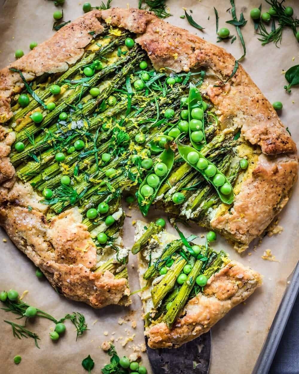 Savory galette topped with asparagus, peas, and herbs, with a slice cut out, showcasing flaky crust and fresh toppings