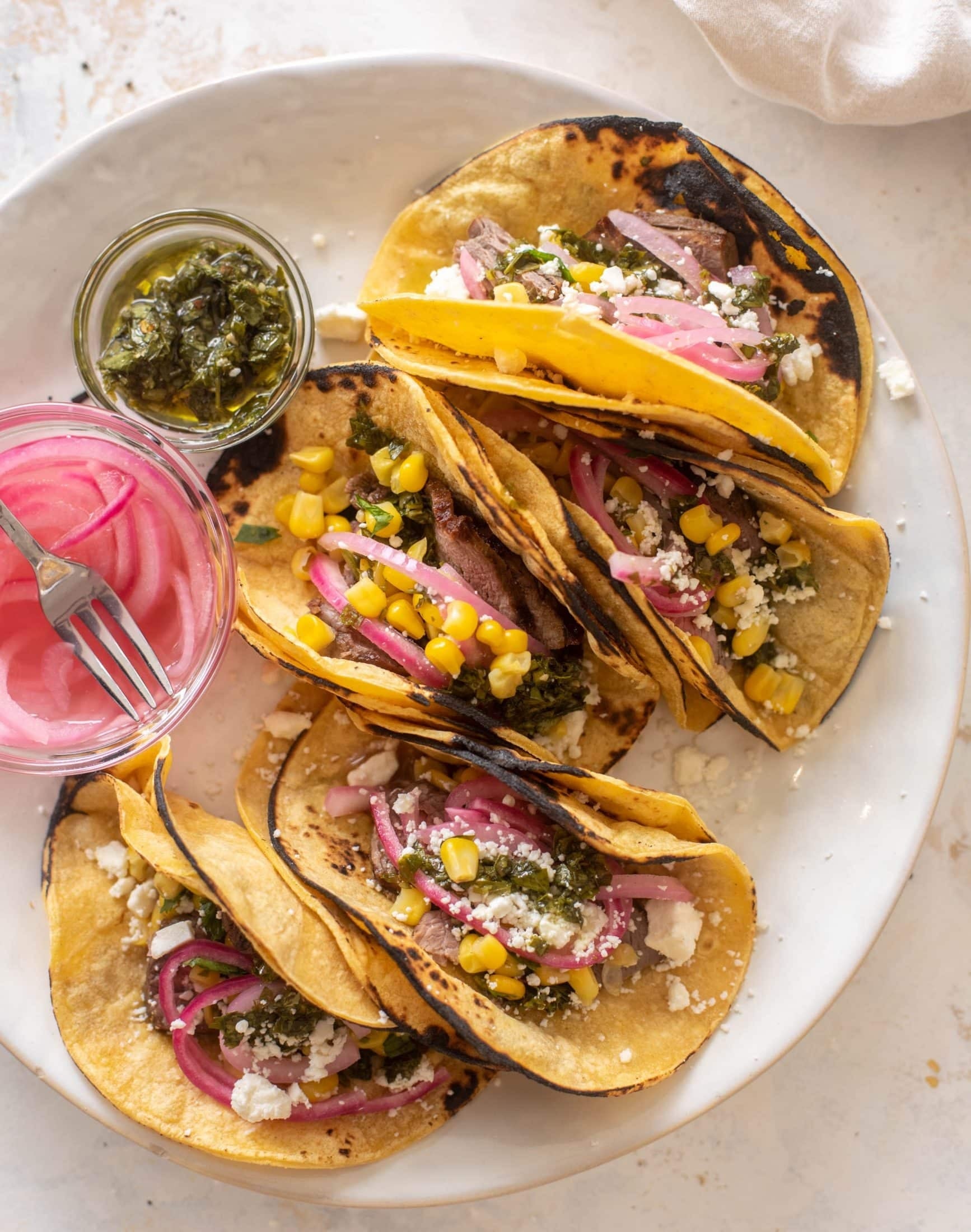Plate of five loaded tacos filled with sliced beef, pickled red onions, corn, cheese, and green salsa. Accompanied by jars of pickled onions and salsa