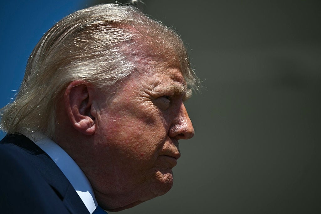 A person in profile, with windswept hair, is outside looking thoughtful, wearing a dark suit and tie