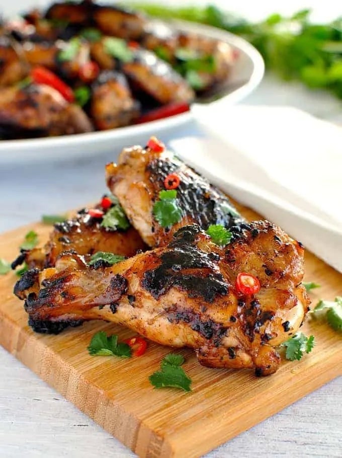 Grilled chicken wings garnished with chili and cilantro on a wooden board, with more wings in the background on a plate