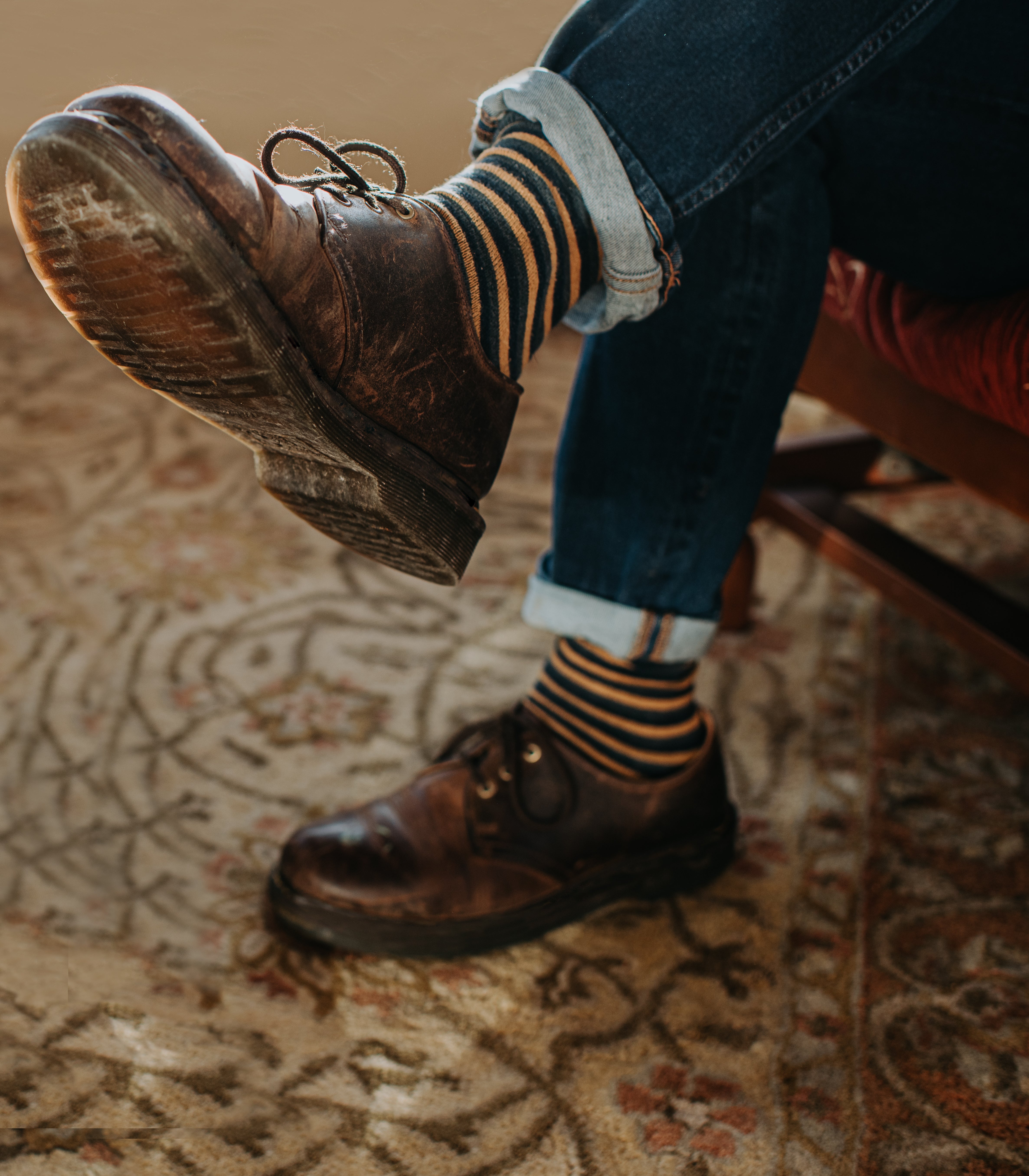 Person wearing worn leather shoes, rolled-up jeans, and striped socks, sitting with 1 limb crossed implicit the different connected a patterned rug