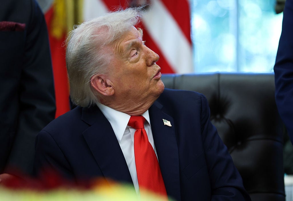 A person in a suit and tie sits in an office setting, looking upward with an attentive expression. An American flag is in the background