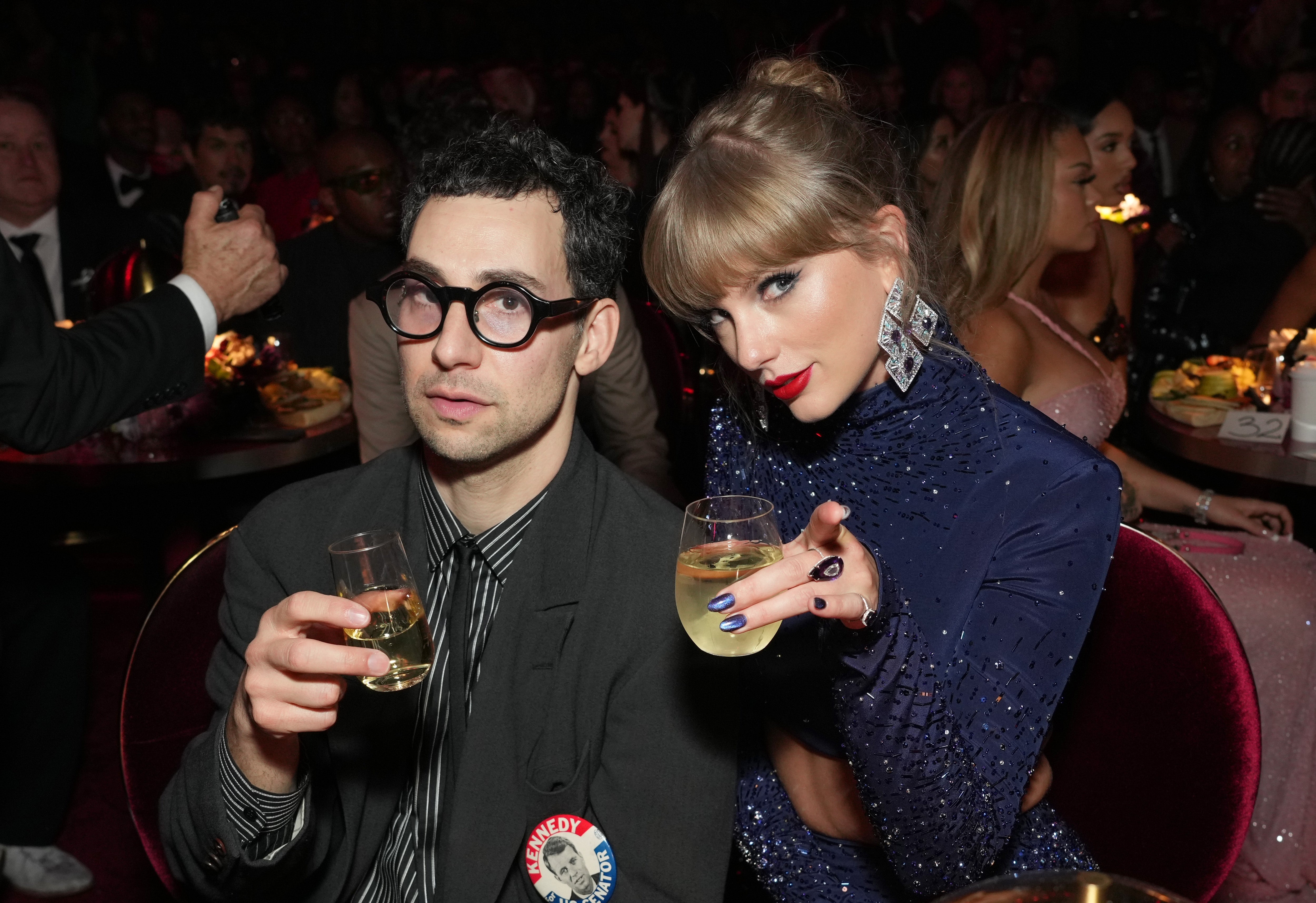 Jack Antonoff in a suit with glasses and Taylor Swift in a sequined outfit hold drinks during an event