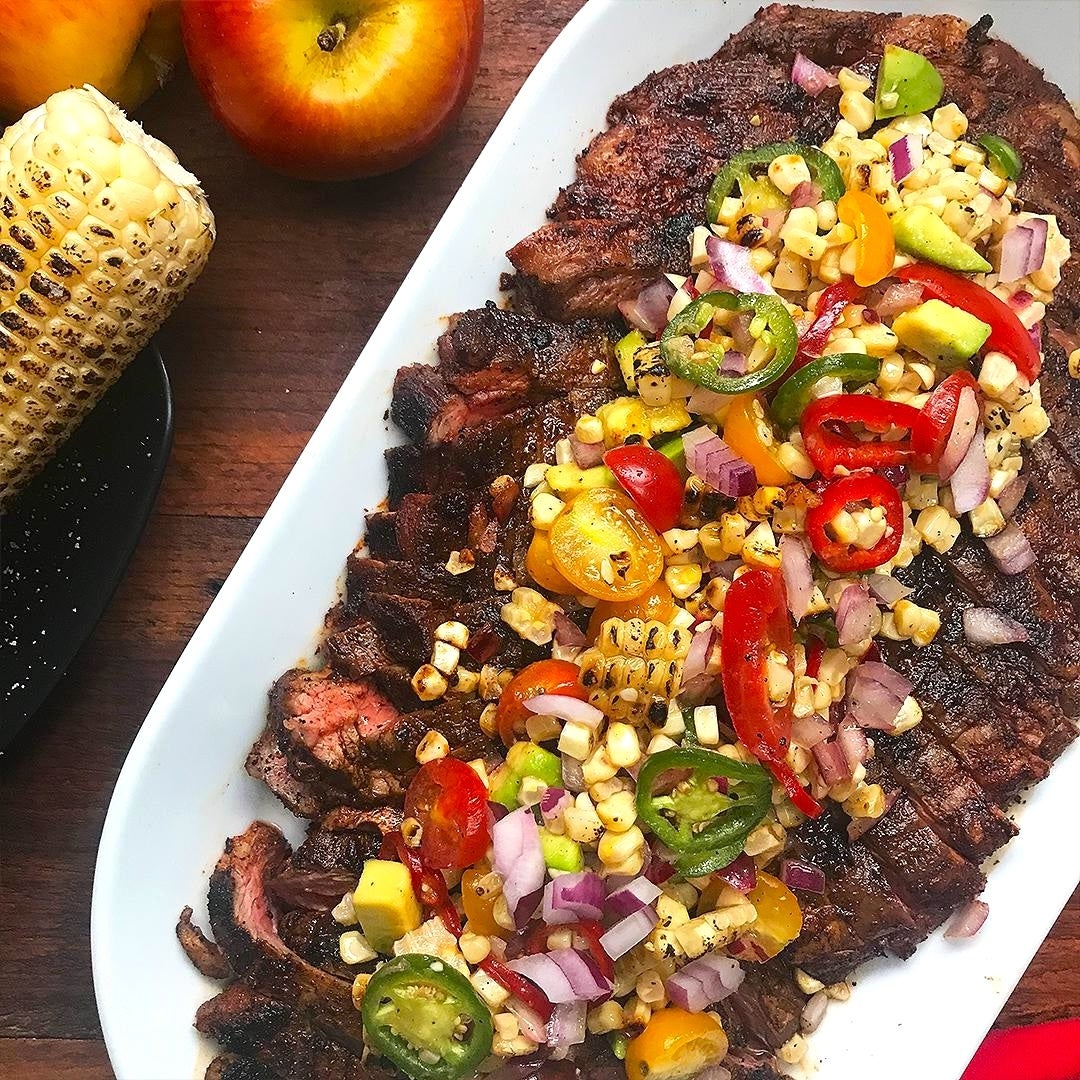 Grilled steak topped with colorful corn salsa on a white platter, surrounded by corn on the cob and apples