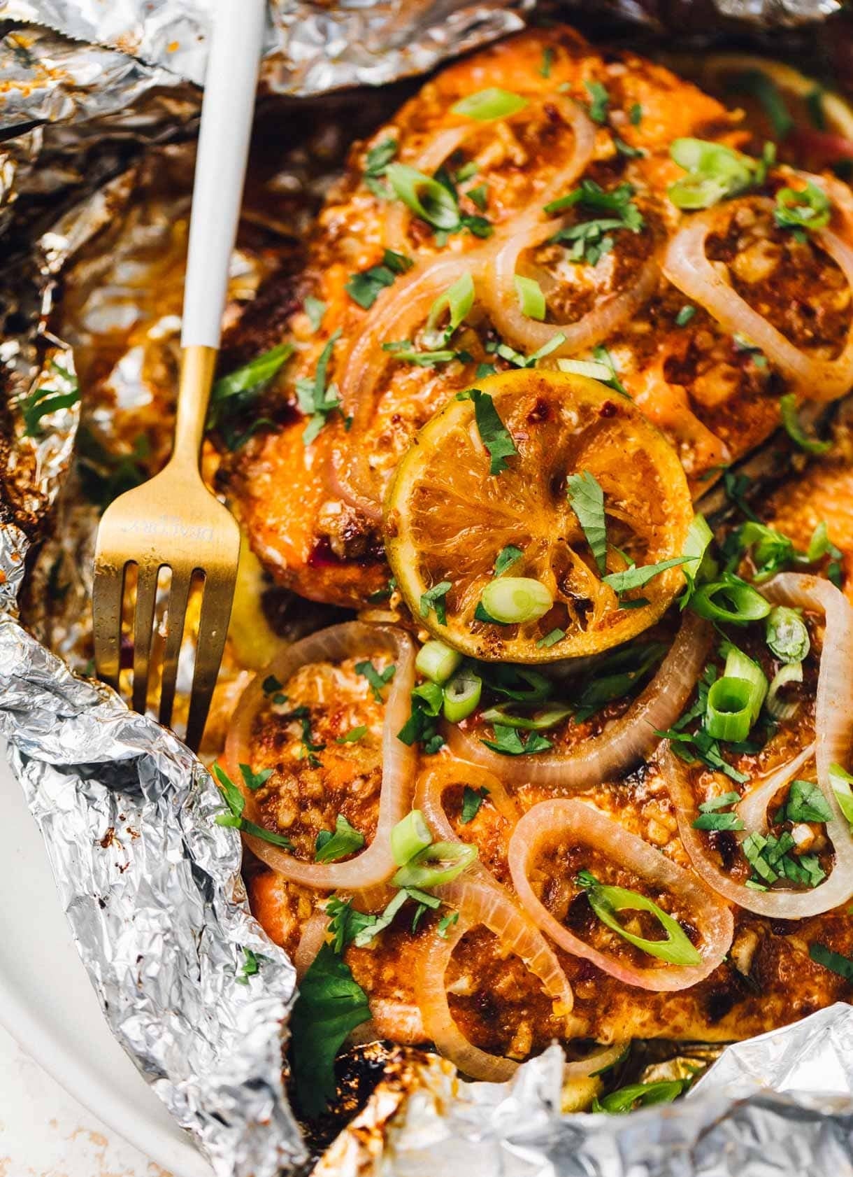 Grilled salmon topped with lemon slices, green onions, and herbs in foil, with a fork on the side
