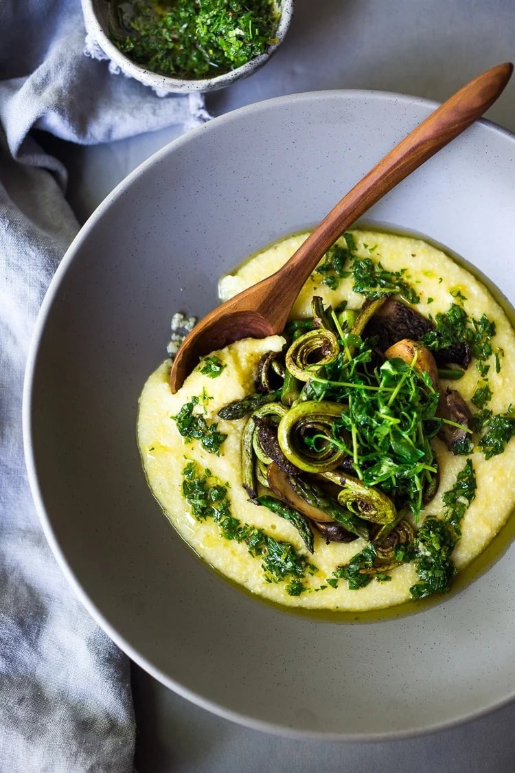 Bowl of creamy polenta topped with sautéed mushrooms, fiddlehead ferns, and fresh herbs drizzled with green sauce