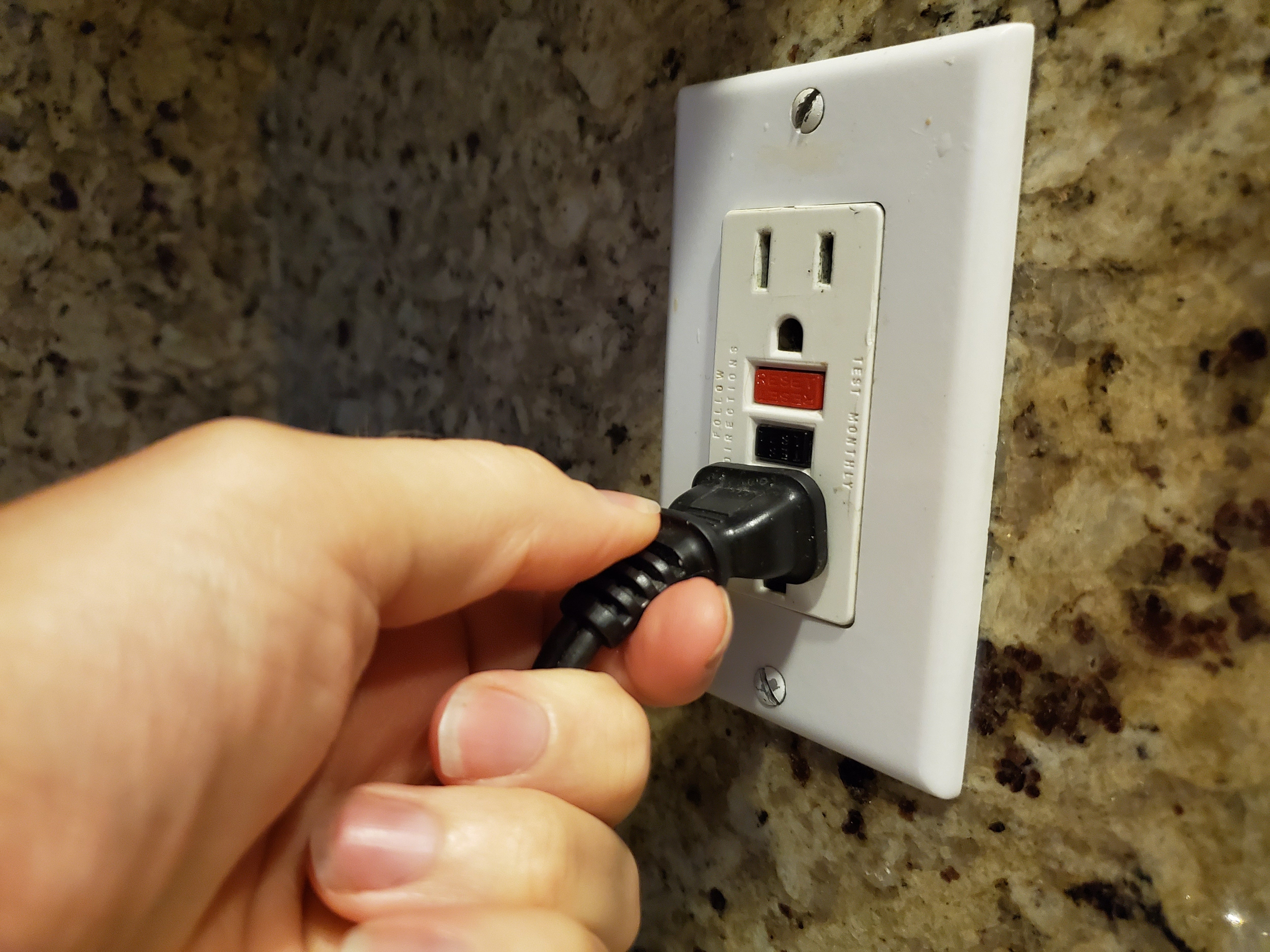 Hand plugging a black electrical cord into a wall outlet with a reset/test button on a granite countertop background