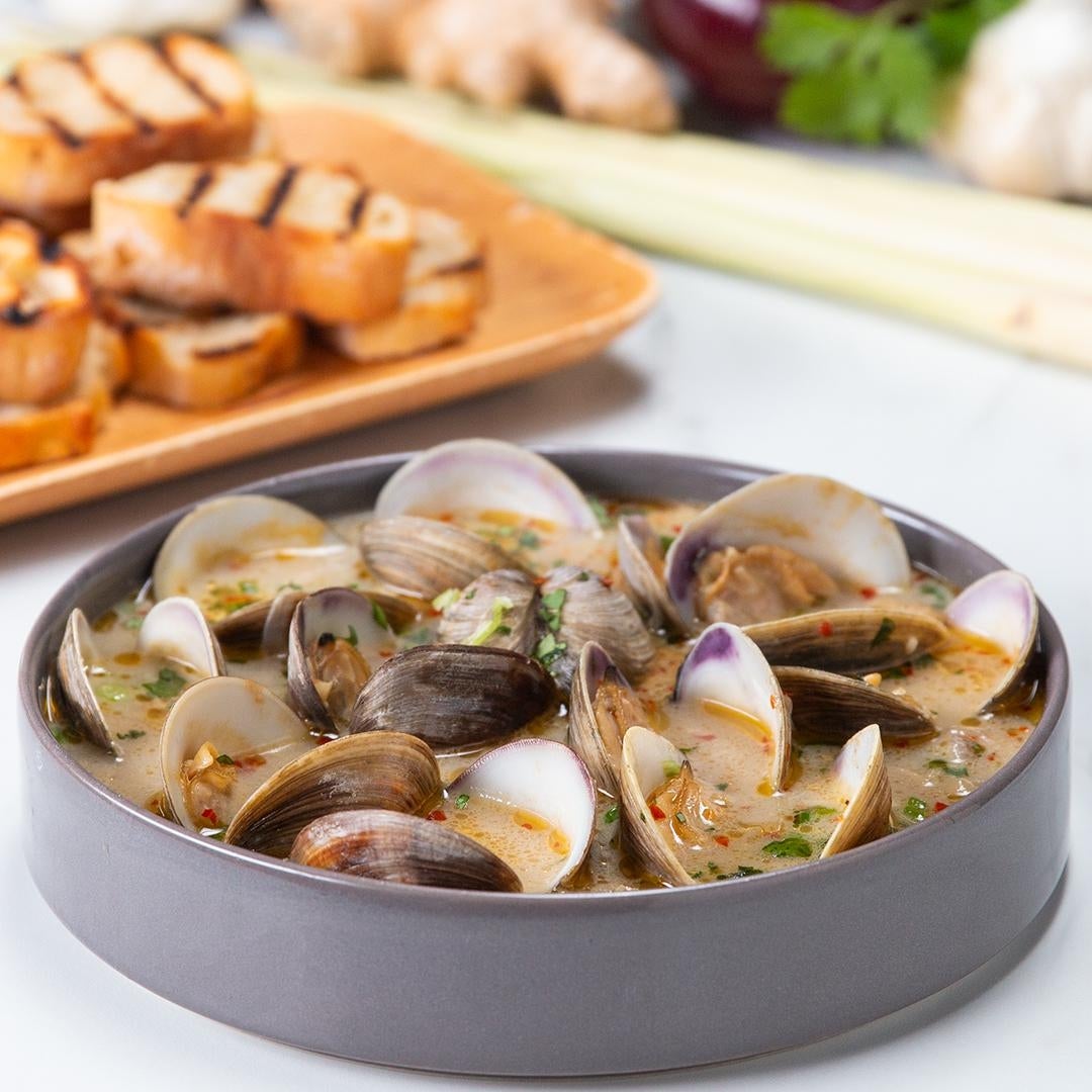 A bowl of clams in a savory broth with herbs, next to grilled bread slices on a plate