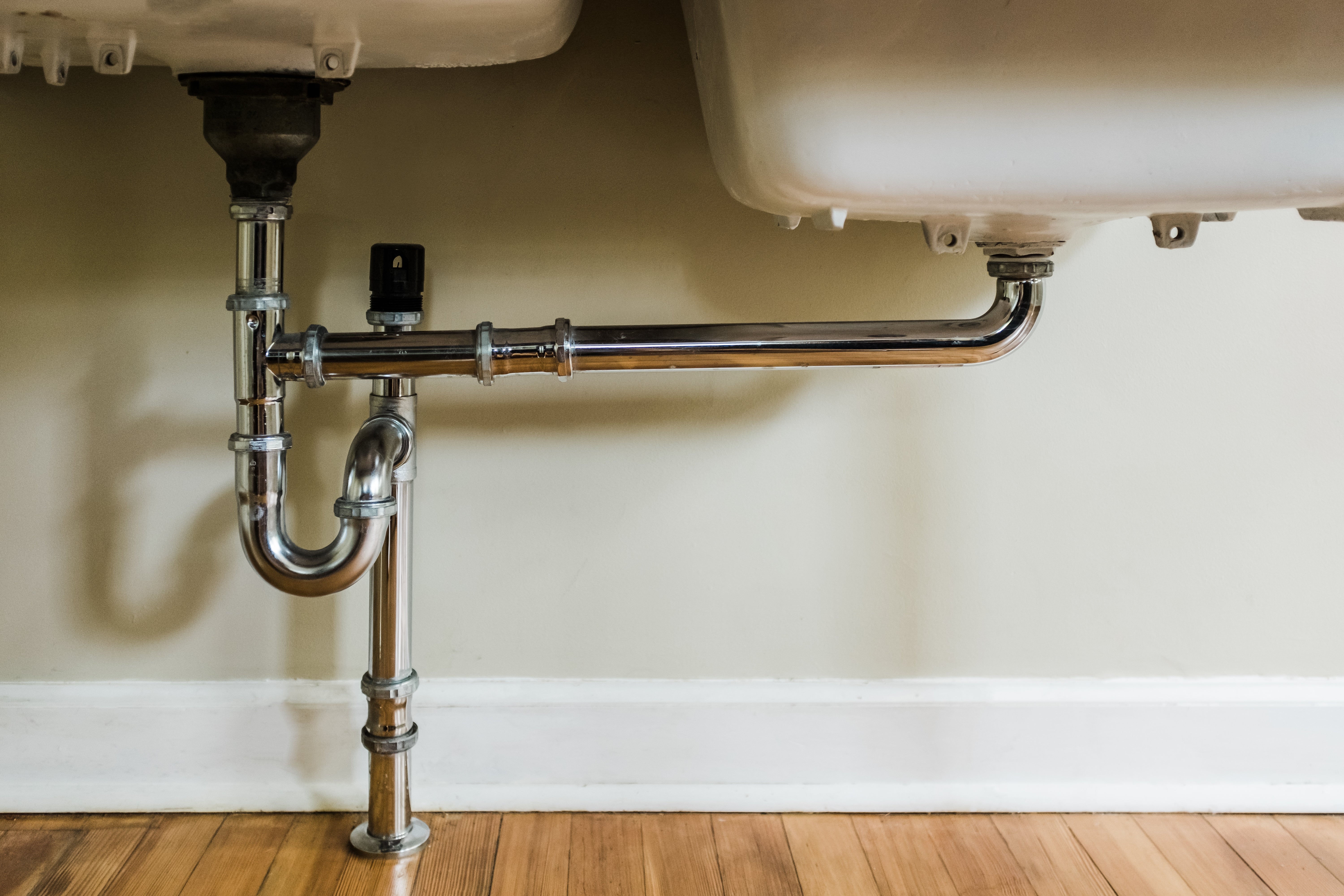 Under-sink plumbing showing chrome pipes and fittings against a beige wall and wooden floor