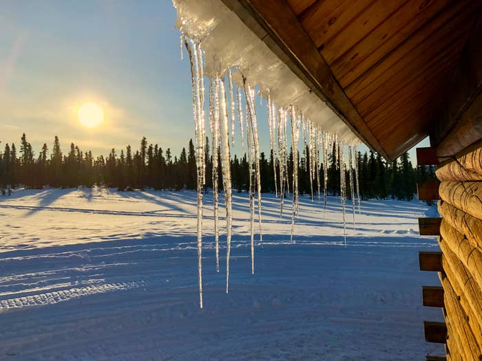 Icicles hang from a cabin roof under a bright sun, with snowy forest landscape in the background