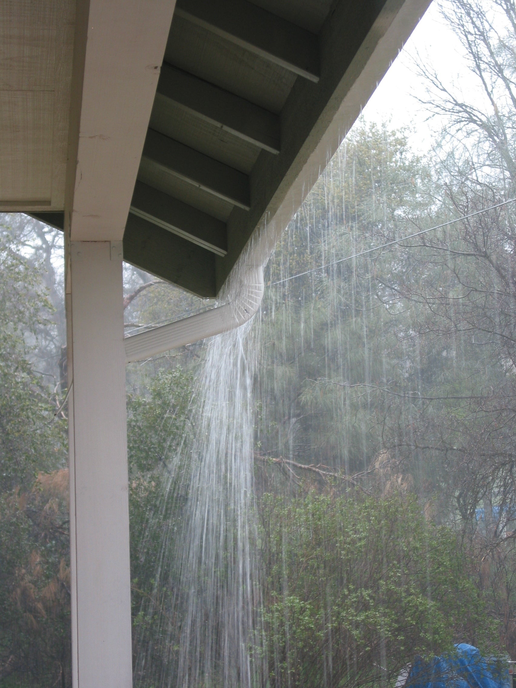 Rainwater is overflowing from a house gutter, cascading down in front of a porch, surrounded by trees and shrubbery