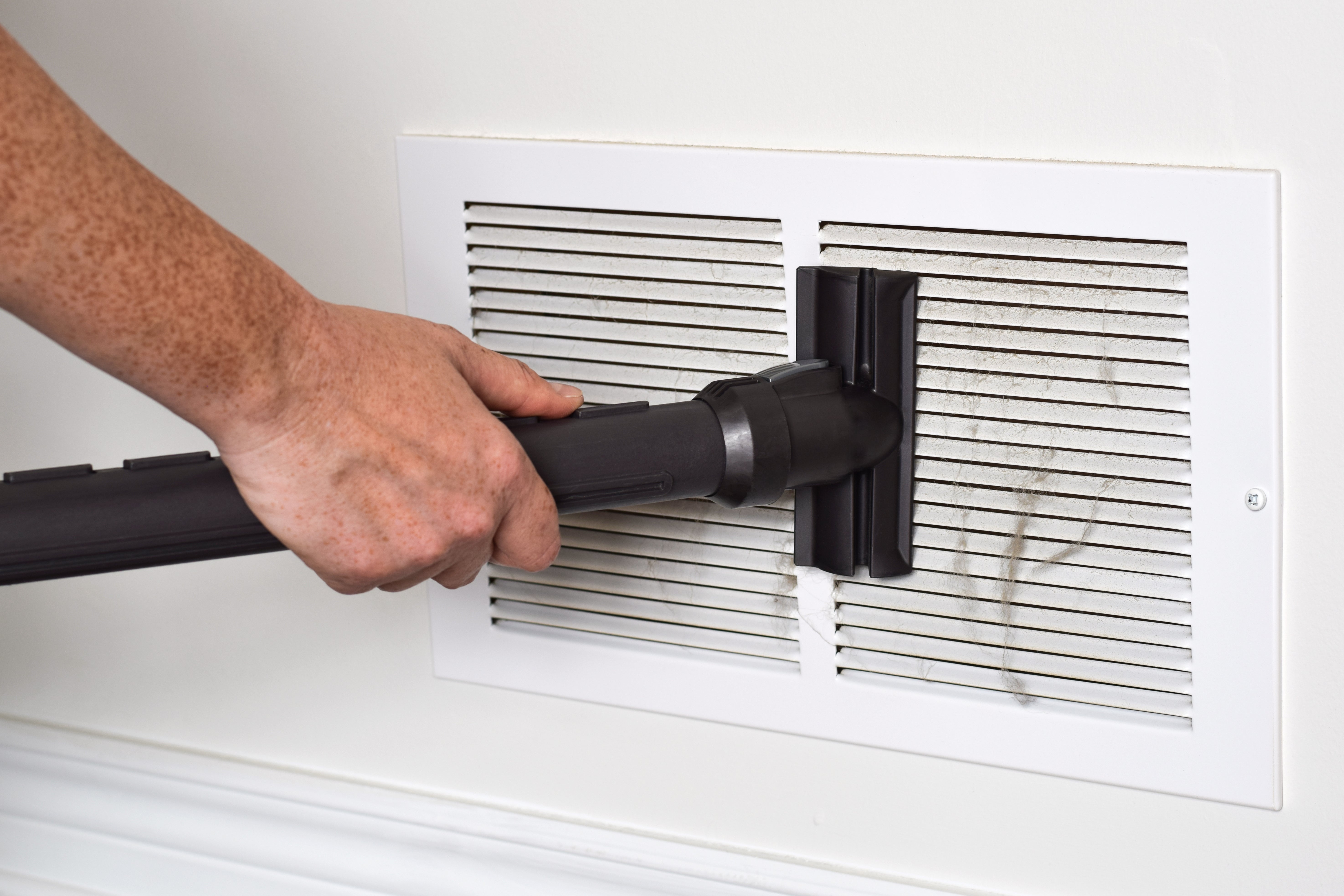 A person uses a vacuum to clean dust from a wall vent indoors
