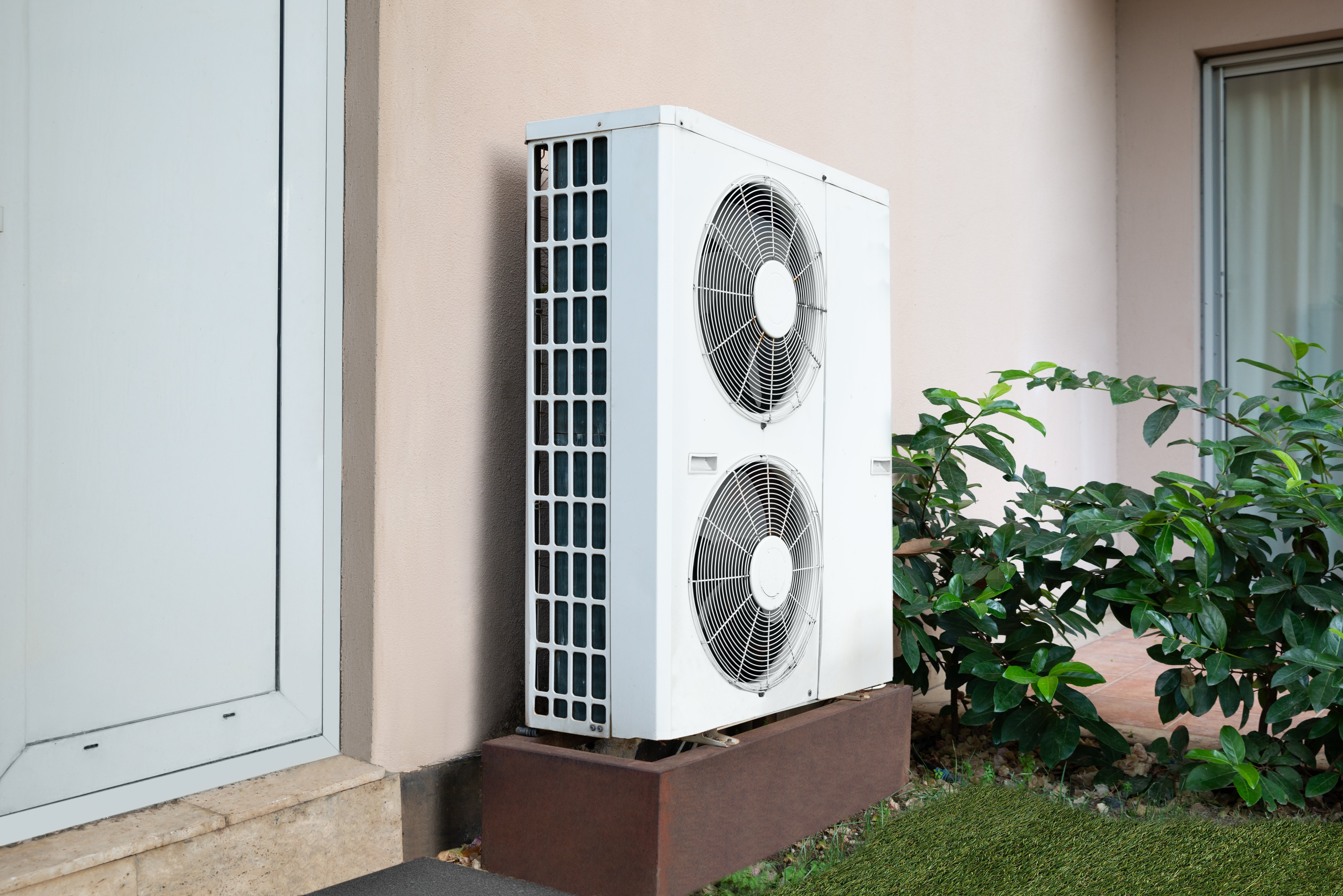 Outdoor air conditioning unit mounted next to a building, surrounded by plants