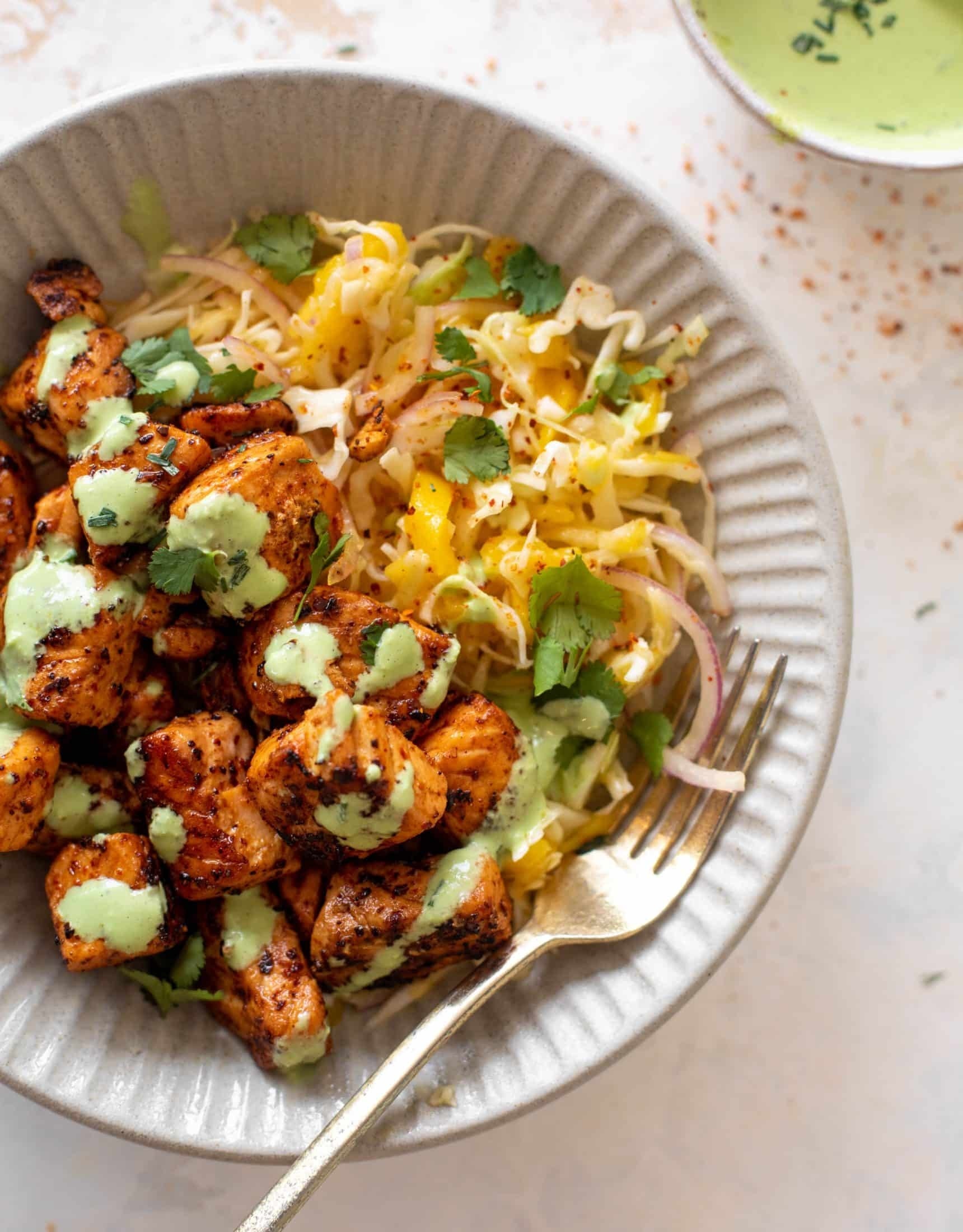 Bowl of seasoned grilled chicken bites topped with green sauce, served with shredded cheese, sliced onions, and cilantro