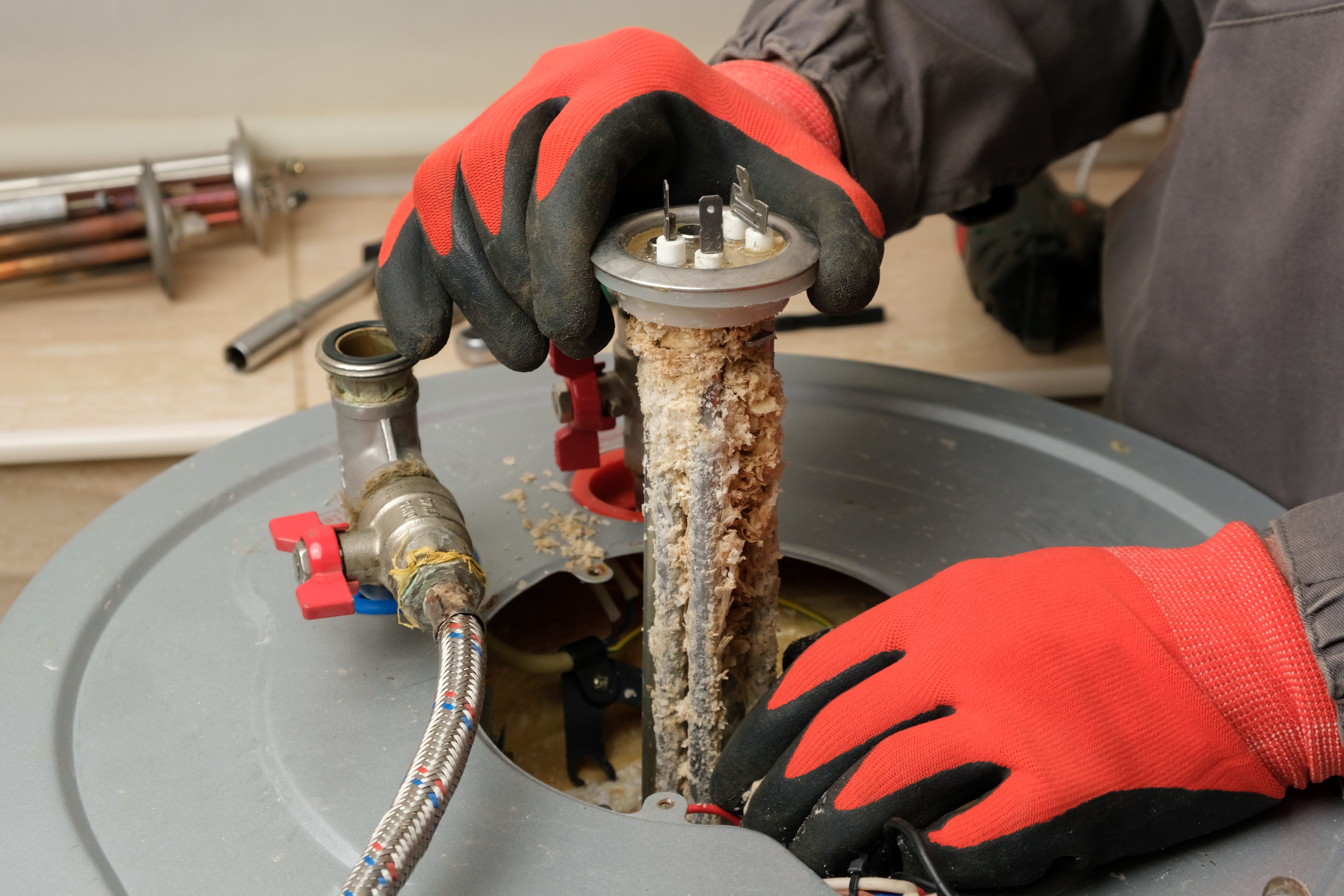 A person wearing red gloves is removing a corroded heating element from a water heater, highlighting maintenance work