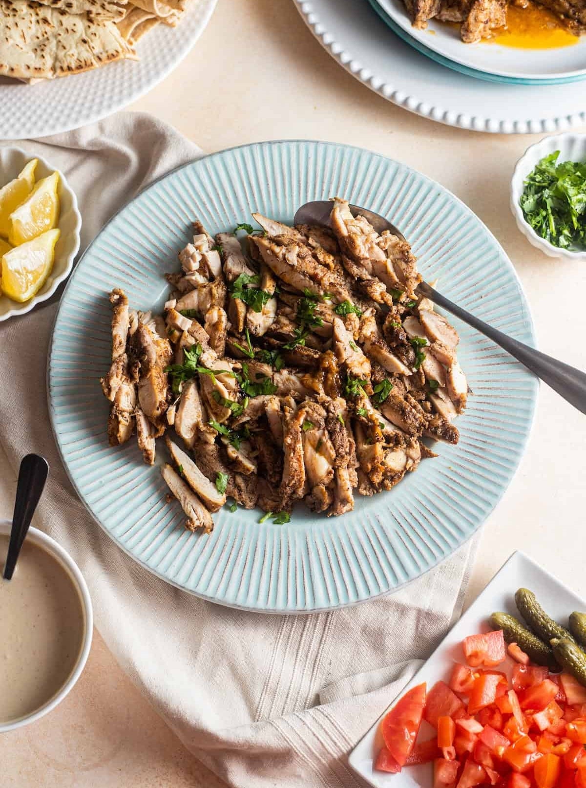 Plate of sliced grilled chicken with herbs, surrounded by lemon wedges, diced tomatoes, small pickles, and sauce on a beige tablecloth