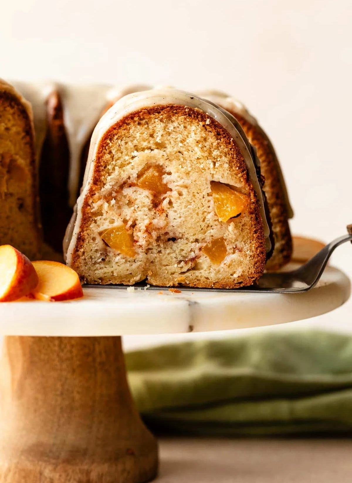 Close-up of a peach bundt cake slice with a creamy glaze, served on a wooden cake stand with sliced peaches beside it