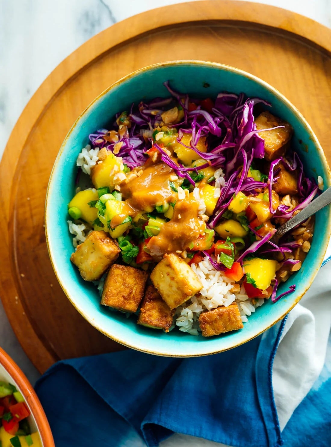 Bowl of rice topped with tofu, red cabbage, mango, and drizzled with sauce, served on a wooden platter with a blue napkin