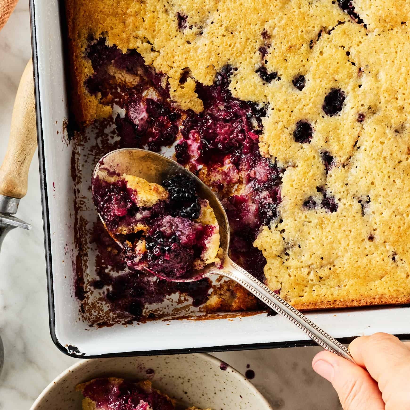 Baked berry cobbler in a pan with a serving spoon scooping a portion; berries and fluffy cake are visible