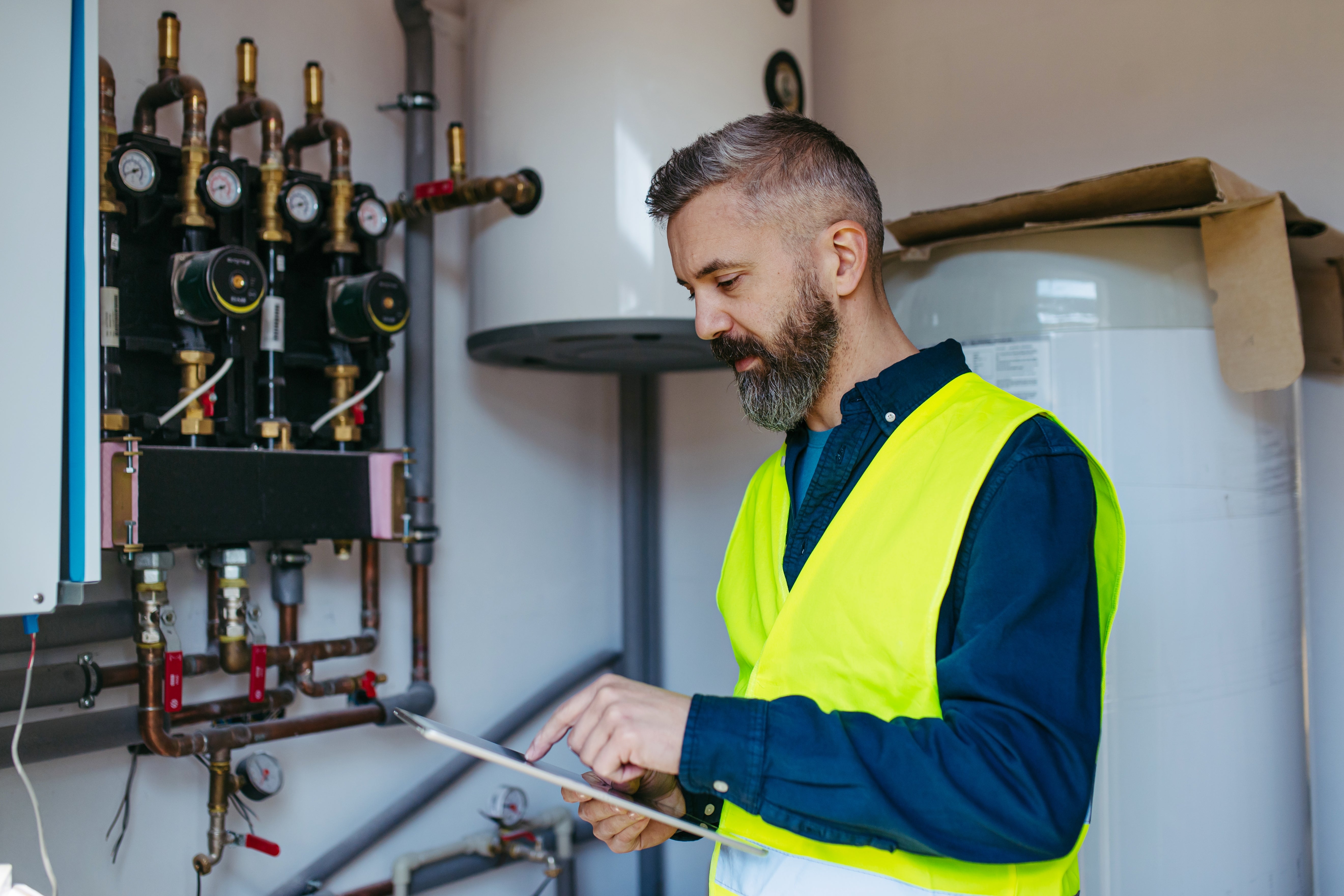 Man with beard in a safety vest inspects machinery in a utility room, using a tablet for notes or data review