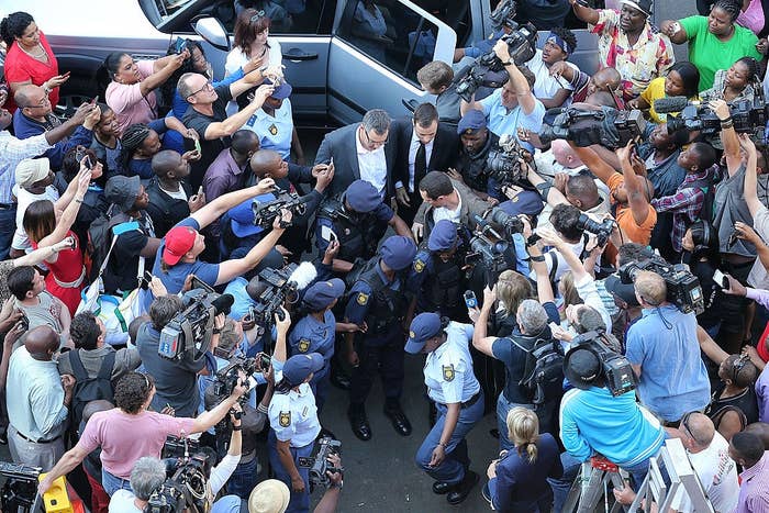Large crowd of reporters and photographers surround security and a public figure exiting a vehicle