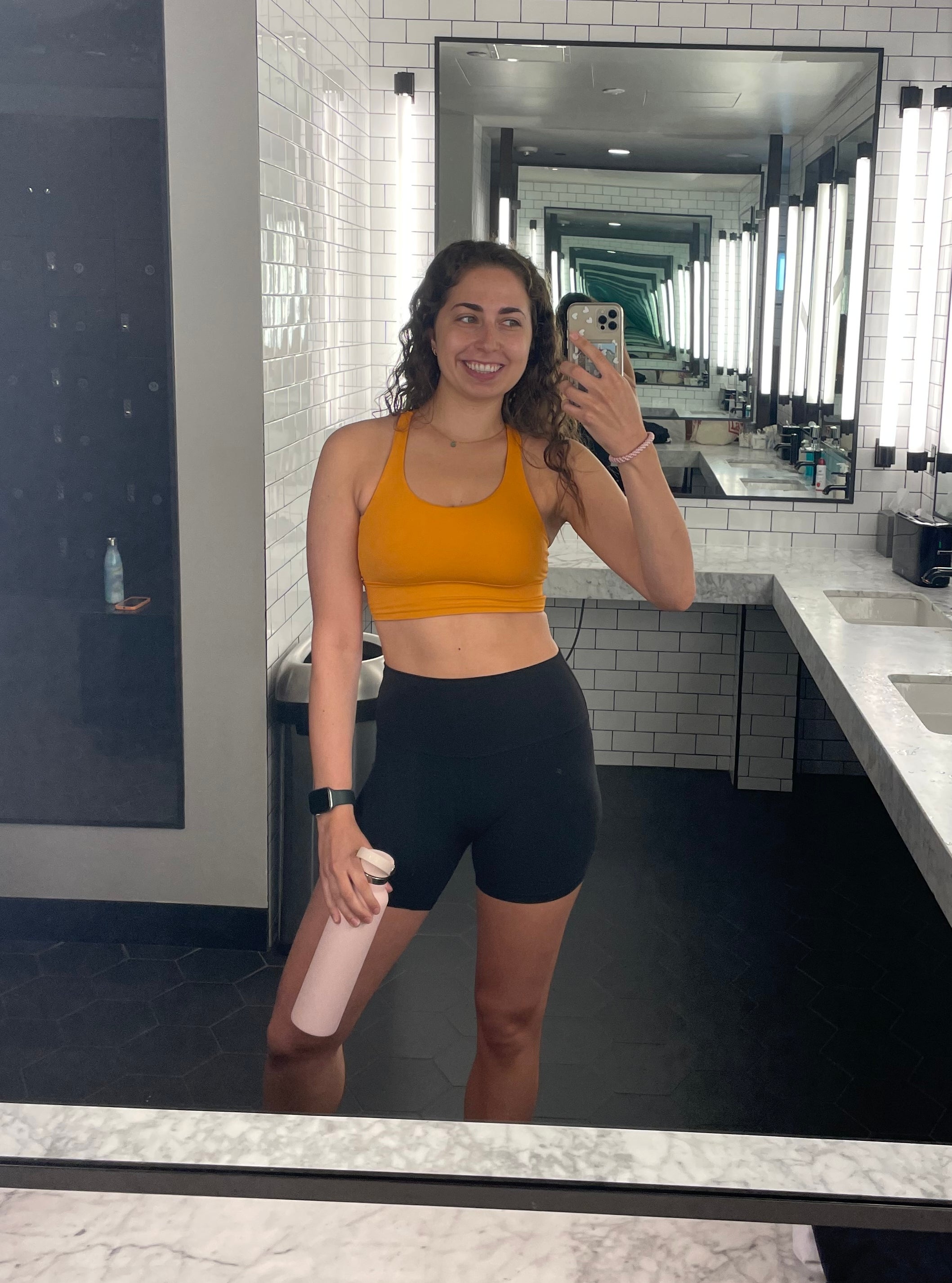 Person tar en speil-selfie på et treningsbad, iført sports-bh og shorts, smilende