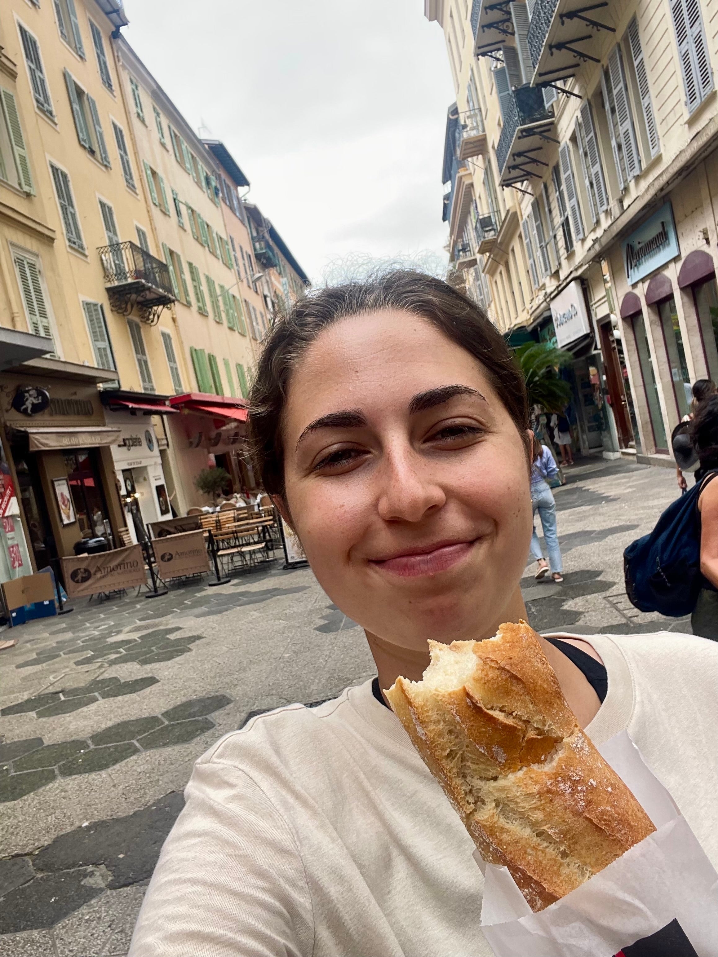 En person som smiler, holder en baguette i en travel bygate med butikker og bygninger i bakgrunnen