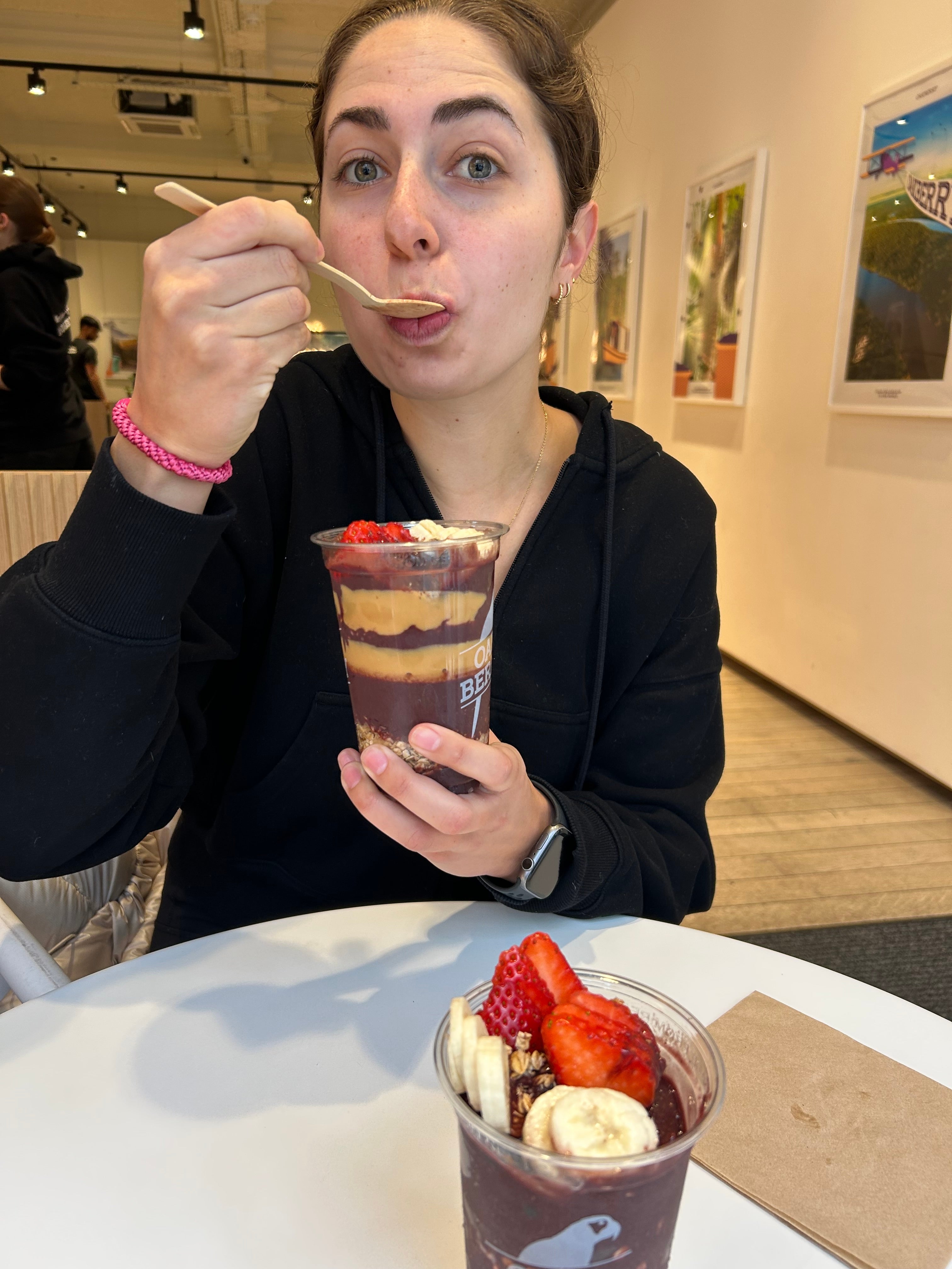 En person på en kafé nyter en acai-skål toppet med bananer og jordbær, iført en uformell svart hettegenser