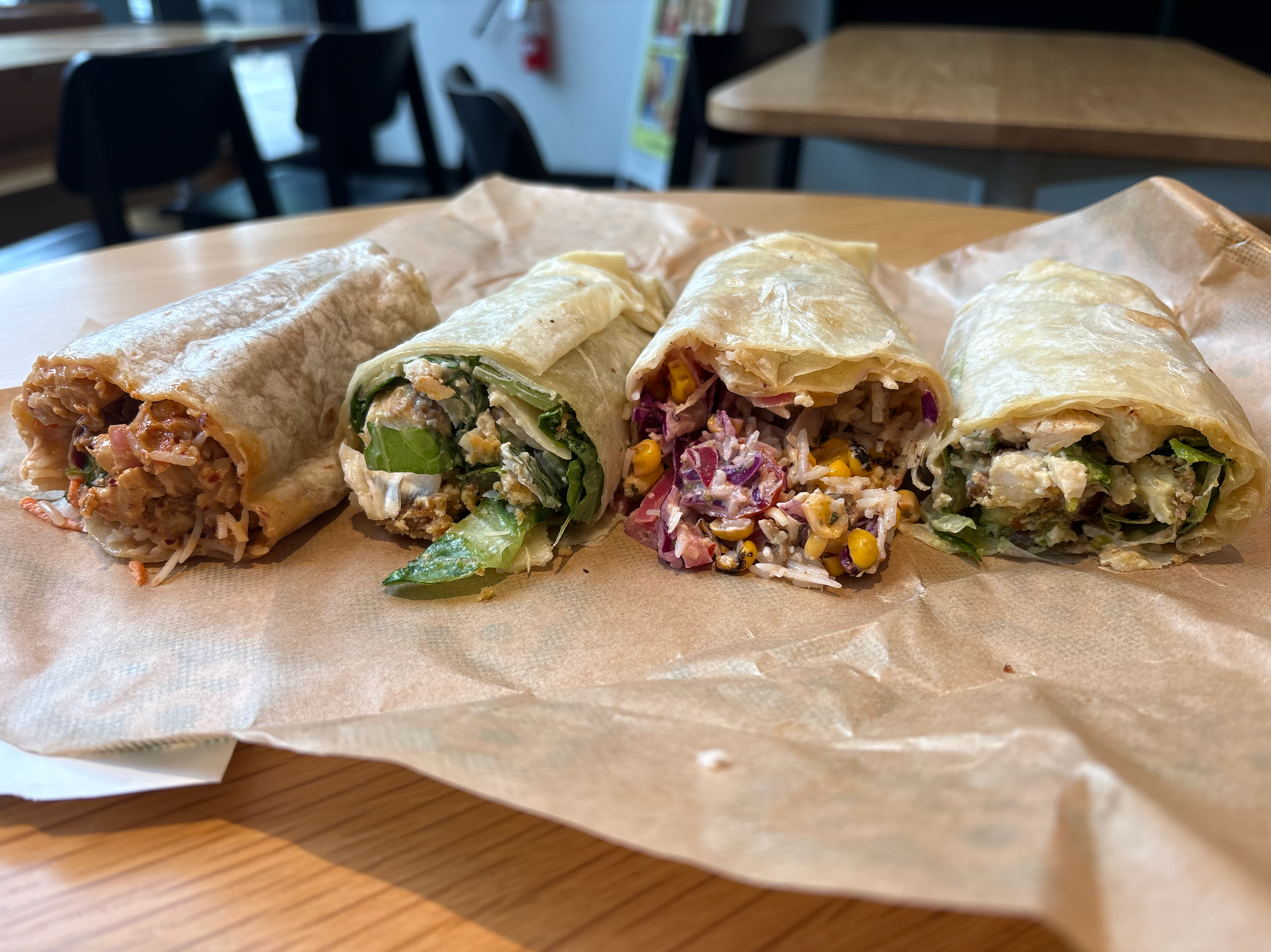 Four different wraps on a brown paper, each cut in half to show diverse fillings like veggies, meats, and grains. They rest on a wooden table