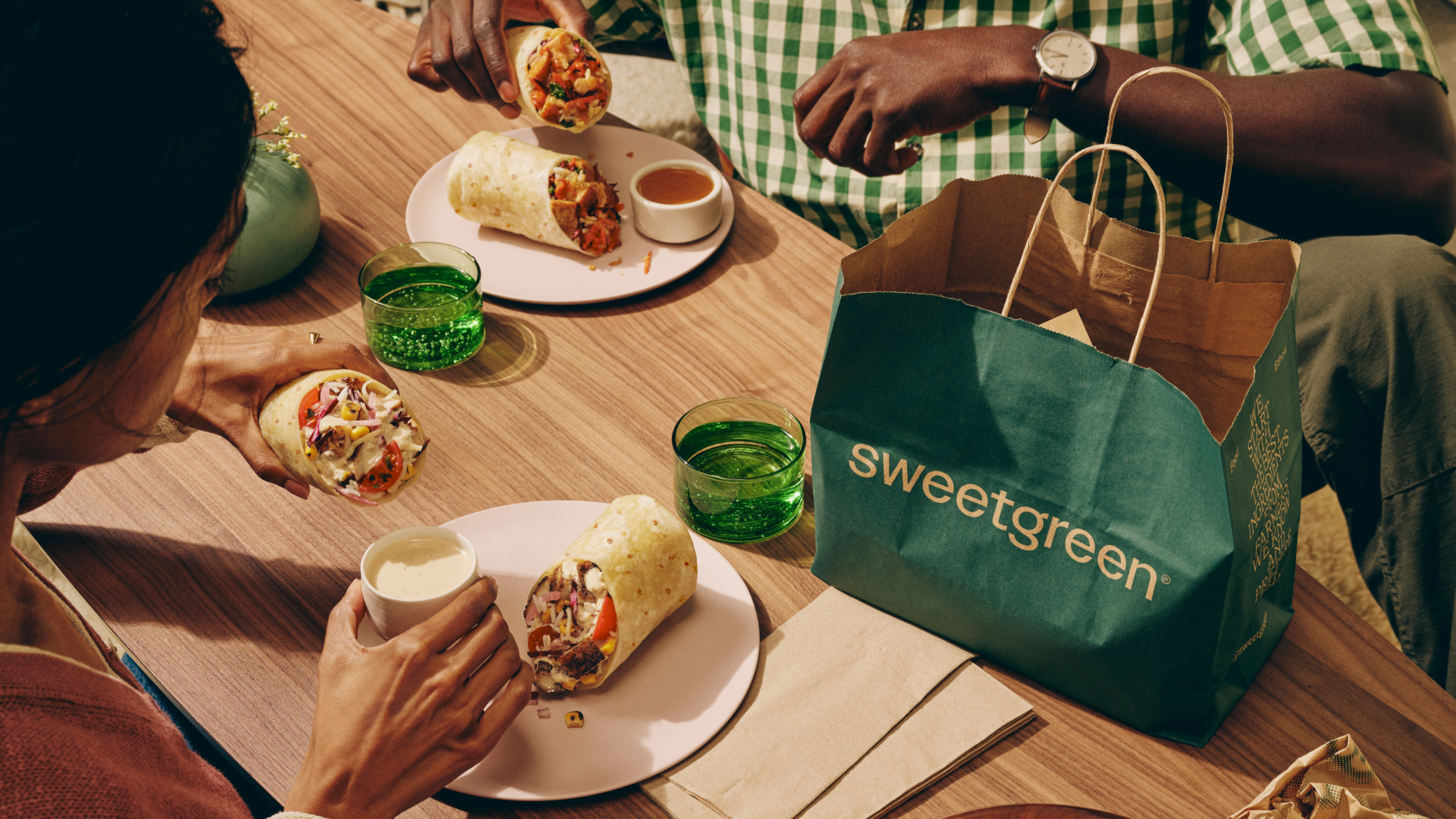 People eating Sweetgreen wraps together with a Sweetgreen paper bag and drinks at a wooden table