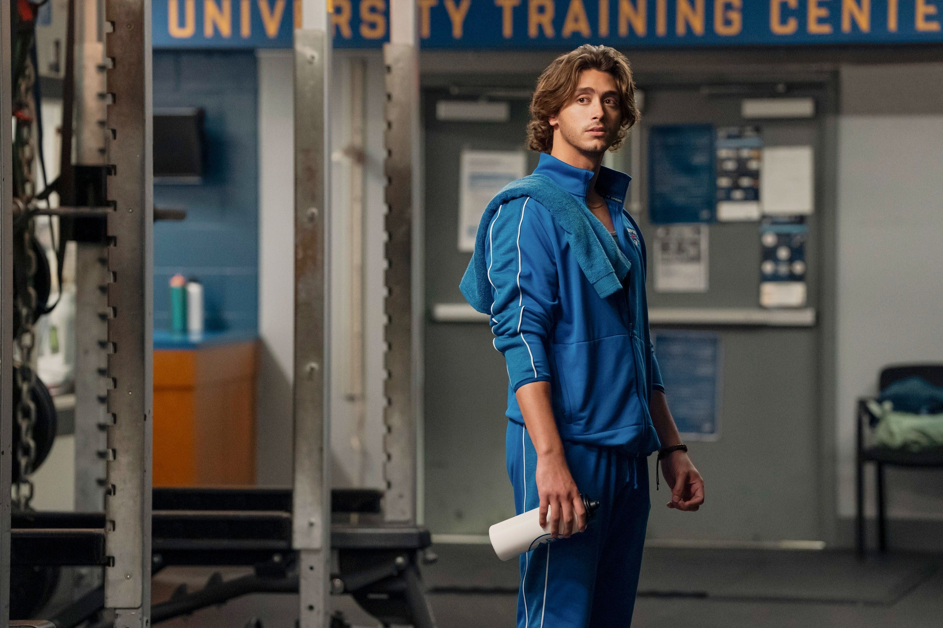 Person in blue sports tracksuit stands in a gym, holding a bottle, with a towel draped over their shoulder
