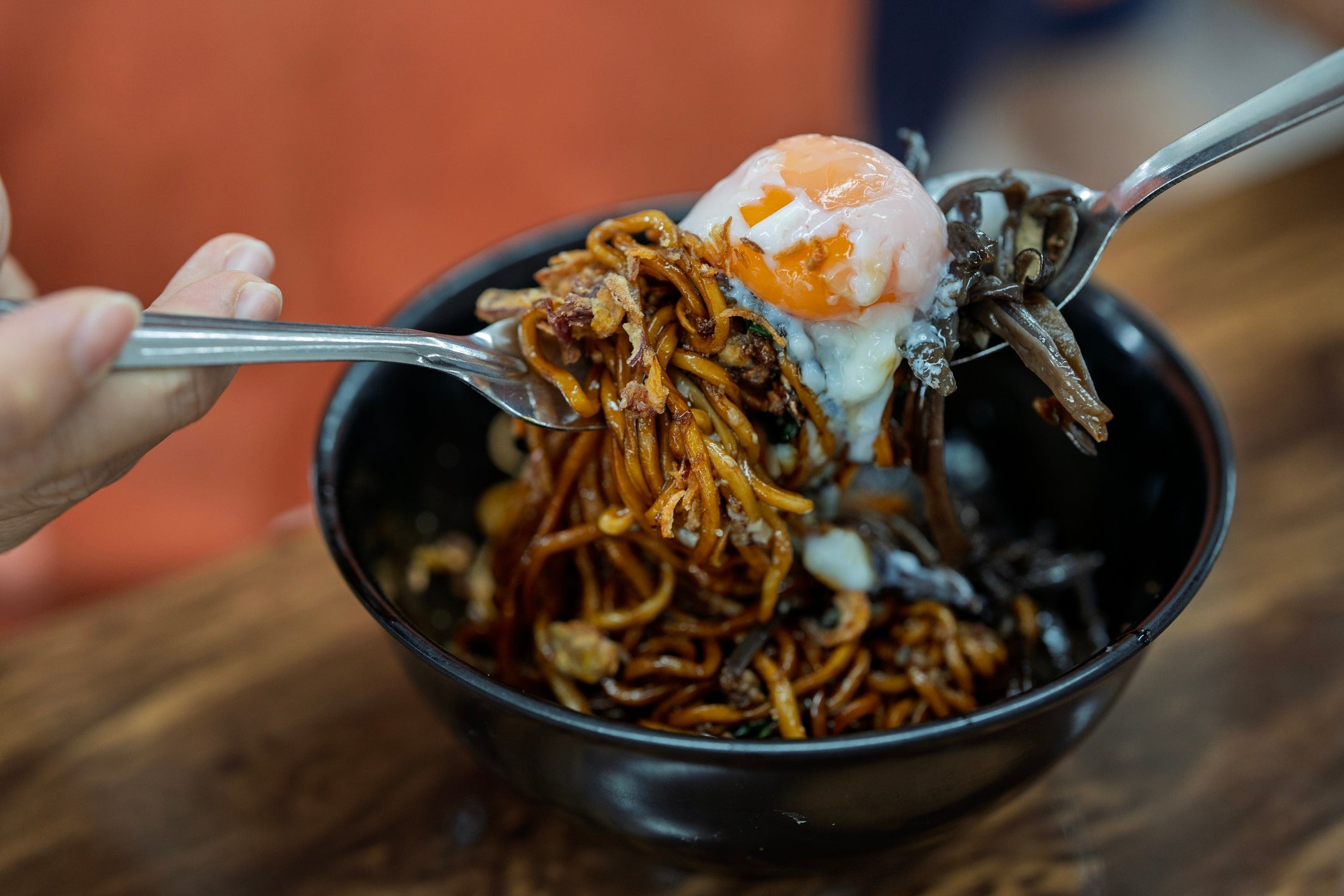 A idiosyncratic holding a forkful of noodles topped with a runny egg, implicit a vessel connected a woody table