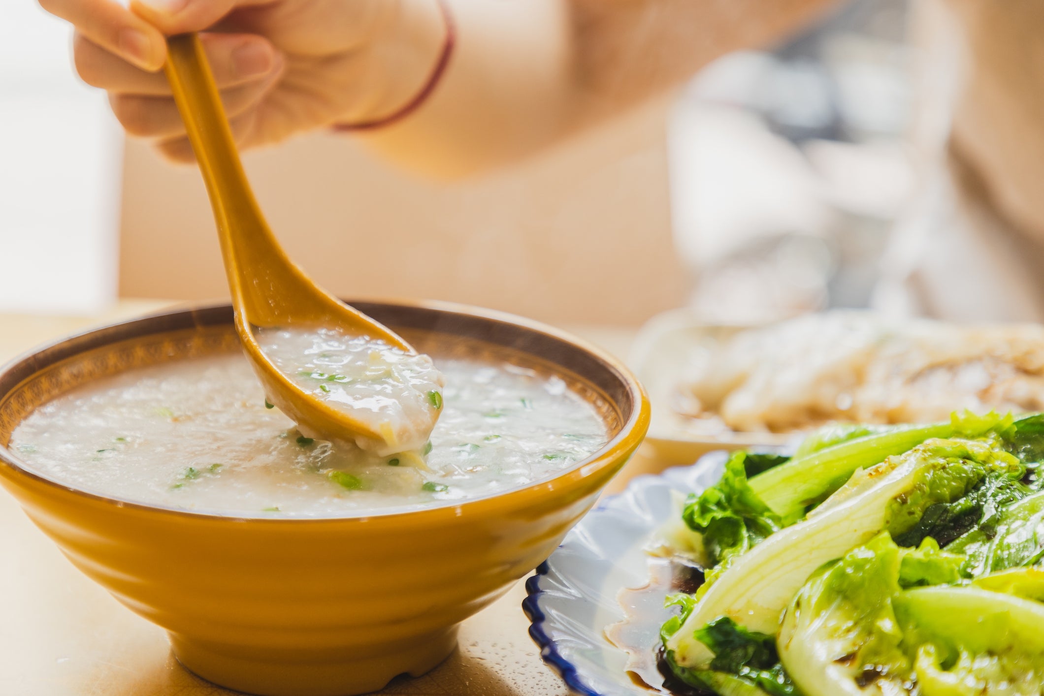 Person serving a vessel of congee with a spoon, garnished with herbs. A sheet of greens is partially disposable nearby