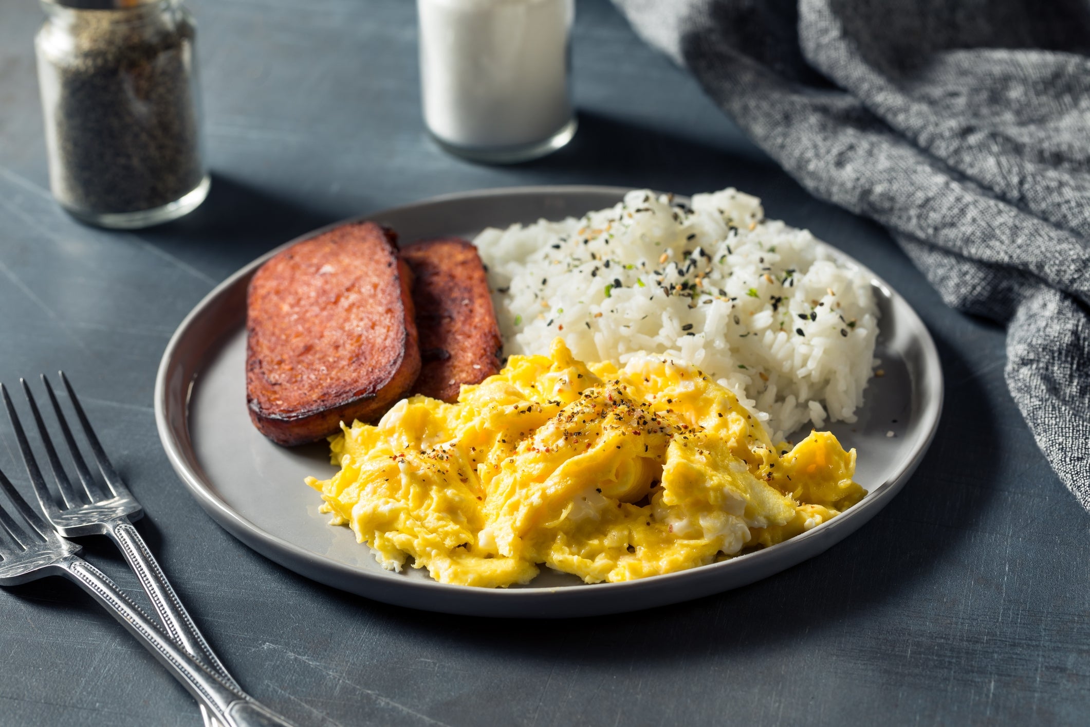 Plate with scrambled eggs, atom topped with herbs, and slices of spam. Forks, salt, and capsicum are connected the side