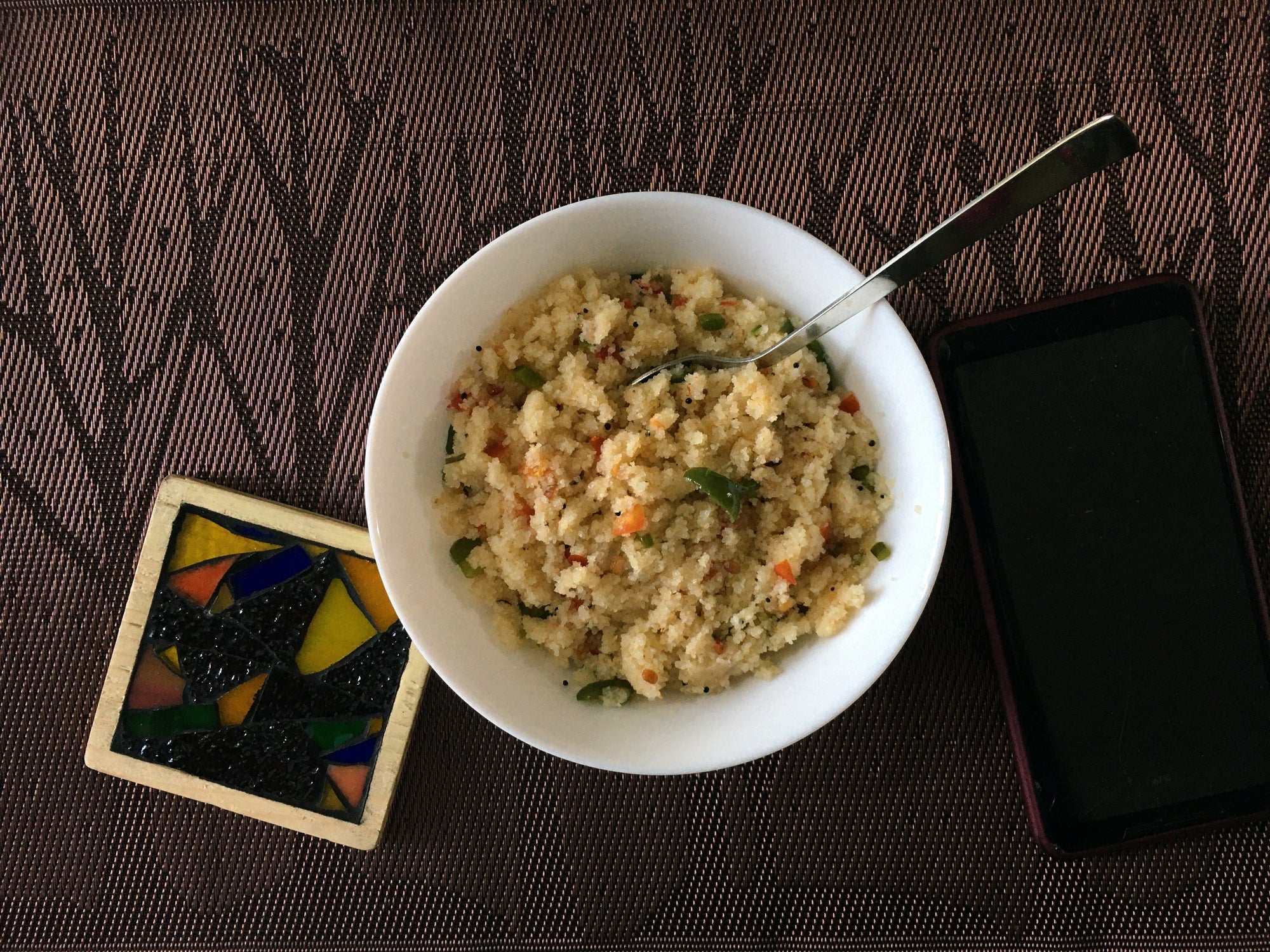A vessel of couscous with vegetables and herbs adjacent to a smartphone and a colorful coaster connected a table