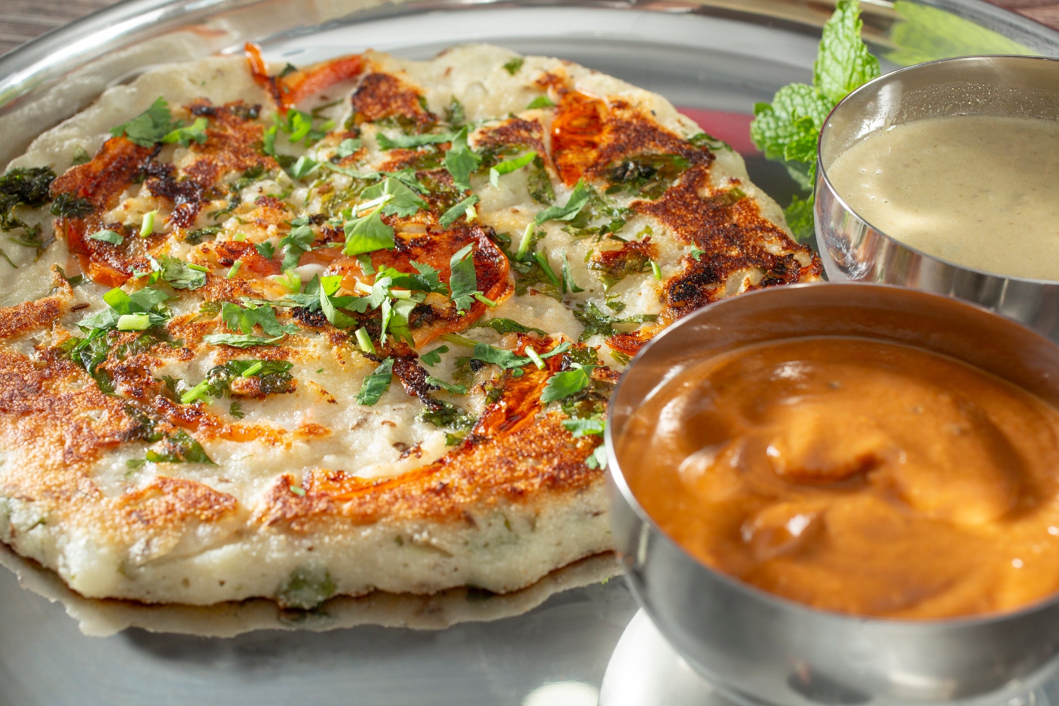 Savory pancake topped with herbs, served with 2 bowls of sauces and a sprig of mint connected a metallic plate