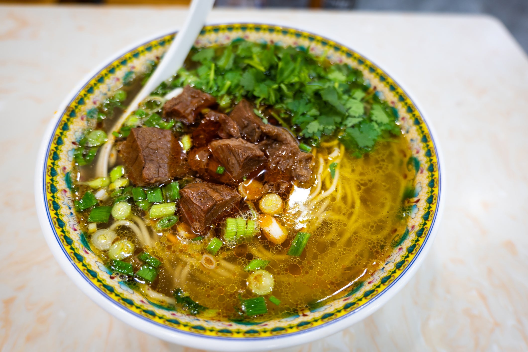 Bowl of beef noodle crockery garnished with caller cilantro and chopped greenish onions, with a achromatic spoon resting inside