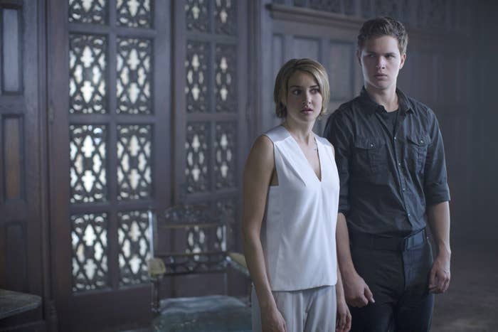 Two actors in a dimly lit room with carved wood details. One wears a sleeveless top, the other a long-sleeved shirt. They look alert and focused