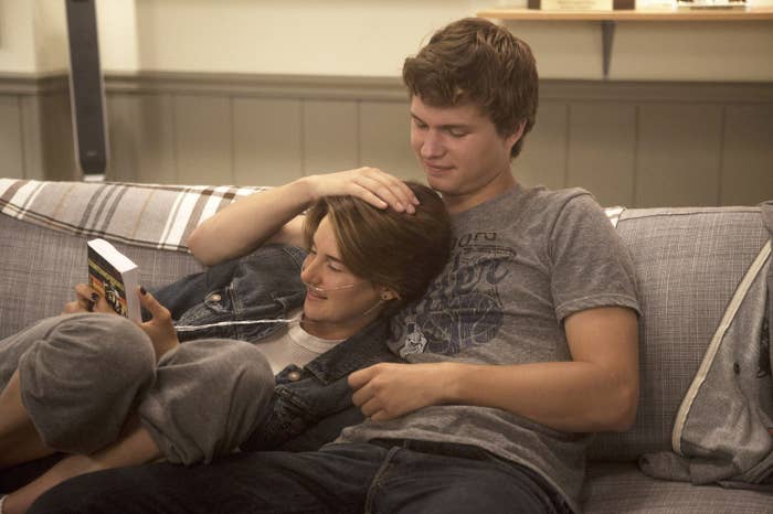 Two people relax on a couch, one reading a book while the other rests their hand on them affectionately, both in casual clothes