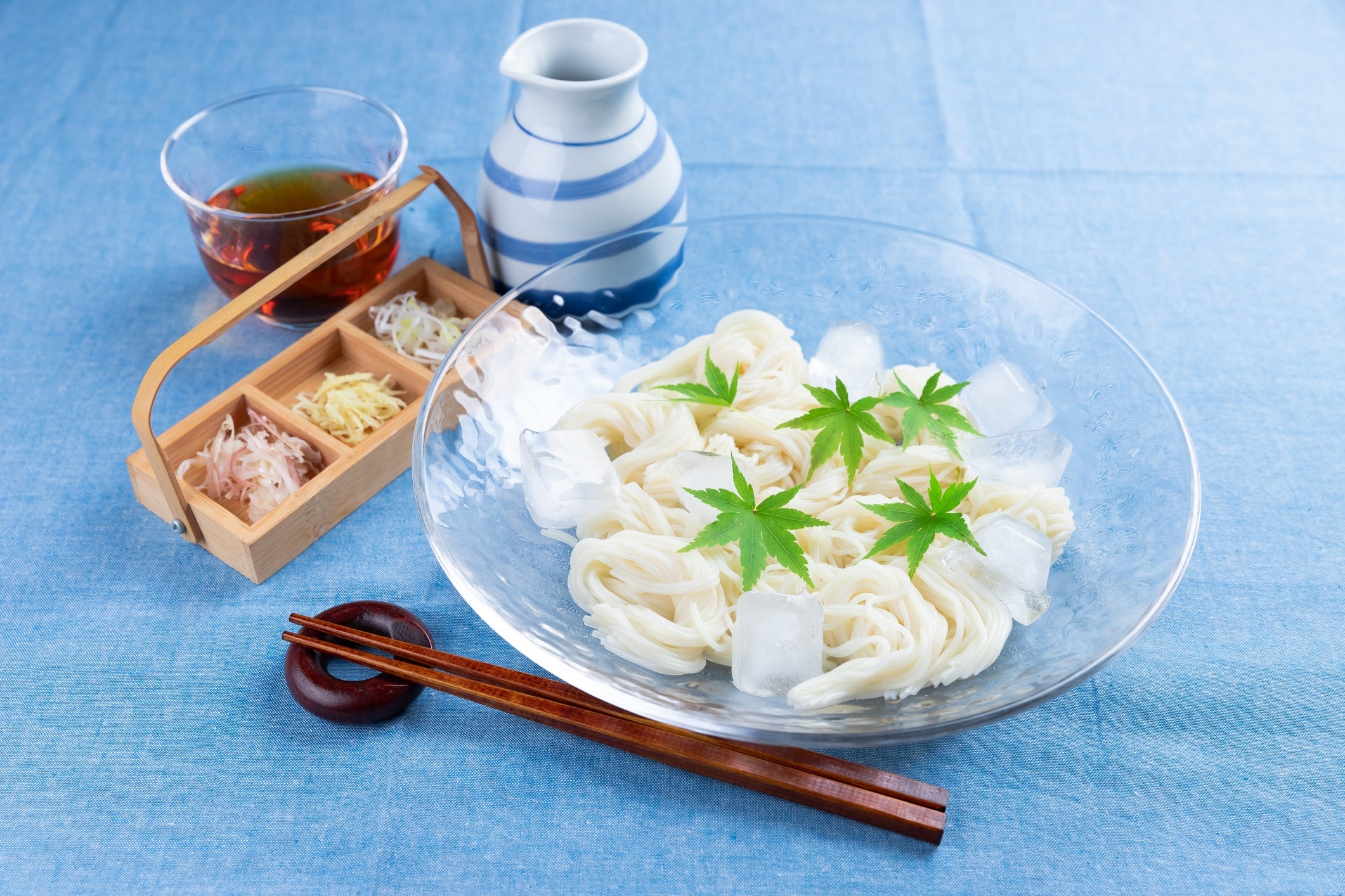 A sheet of chilled noodles with crystal cubes and garnished with greenish leaves, served with dipping condiment and condiments connected the side