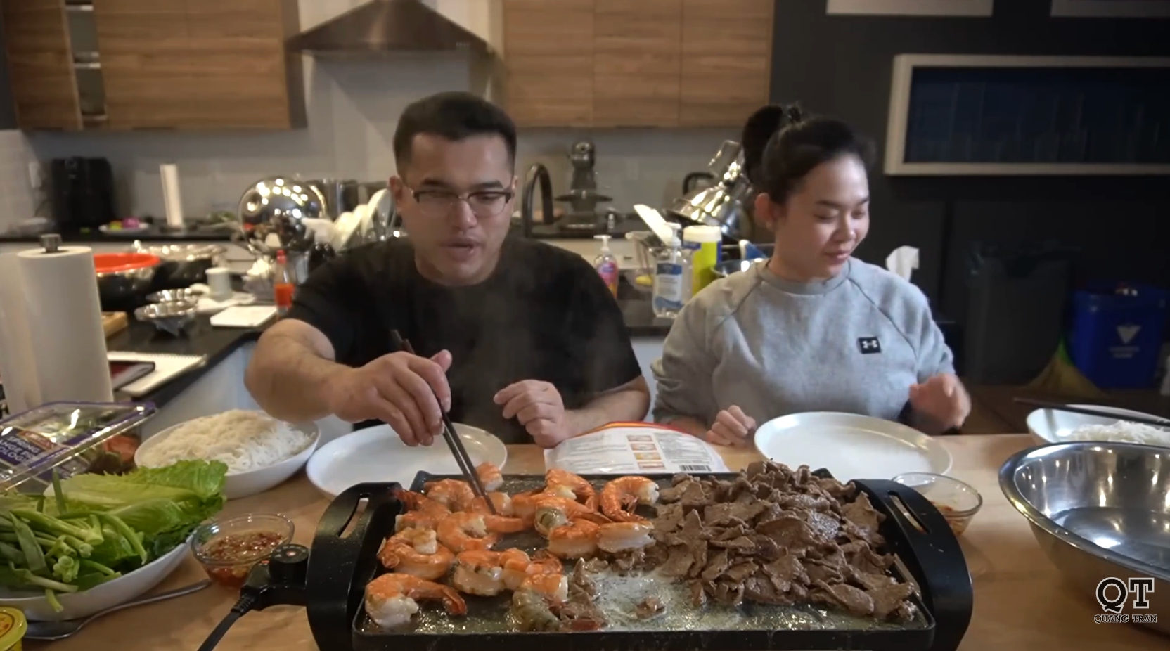 Two radical cooking connected a tabletop grill with shrimp and meat. Various ingredients and cooking utensils are dispersed retired connected the room counter