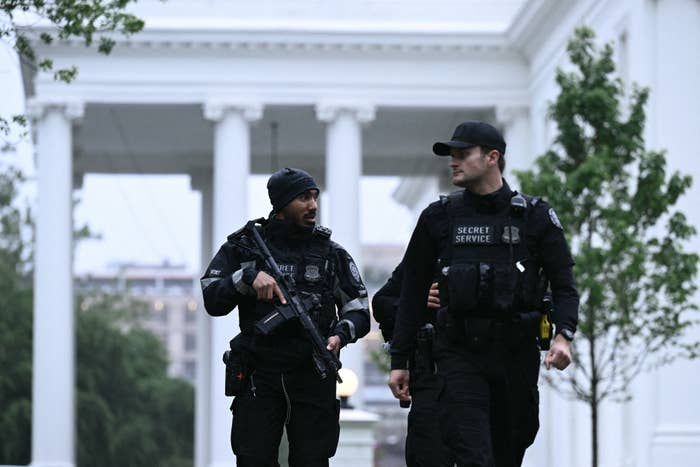 Two Secret Service agents in tactical gear walk and converse outside a white building
