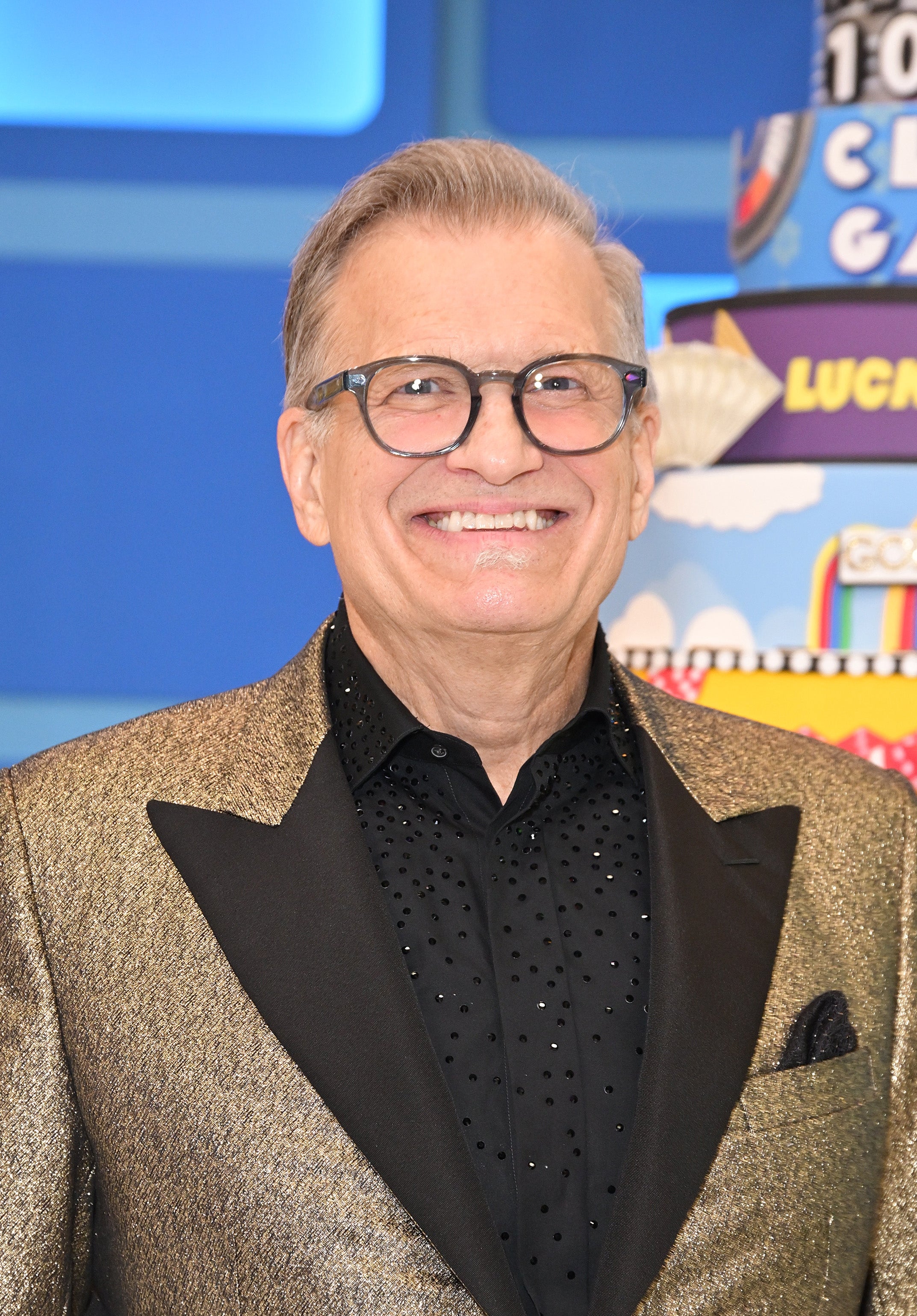A person wearing glasses, a patterned shirt, and a glittery blazer smiles in front of a colorful background