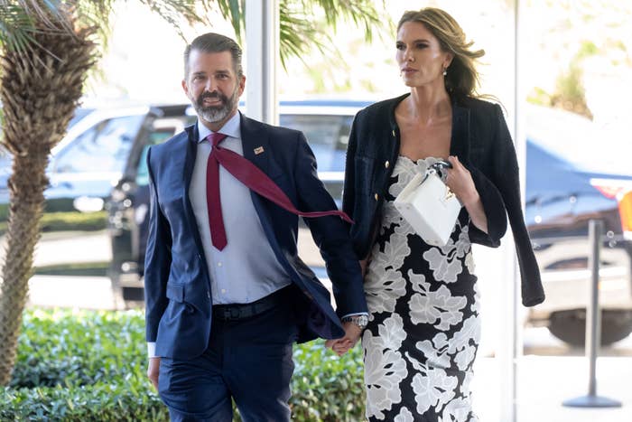 Man in a suit with loose tie and woman in a floral dress and jacket walking outdoors, holding hands