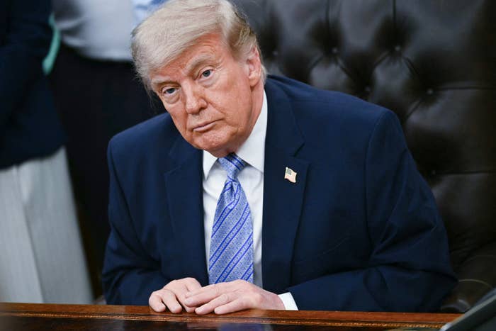A person in a suit and tie sits at a desk, looking at the camera with a serious expression