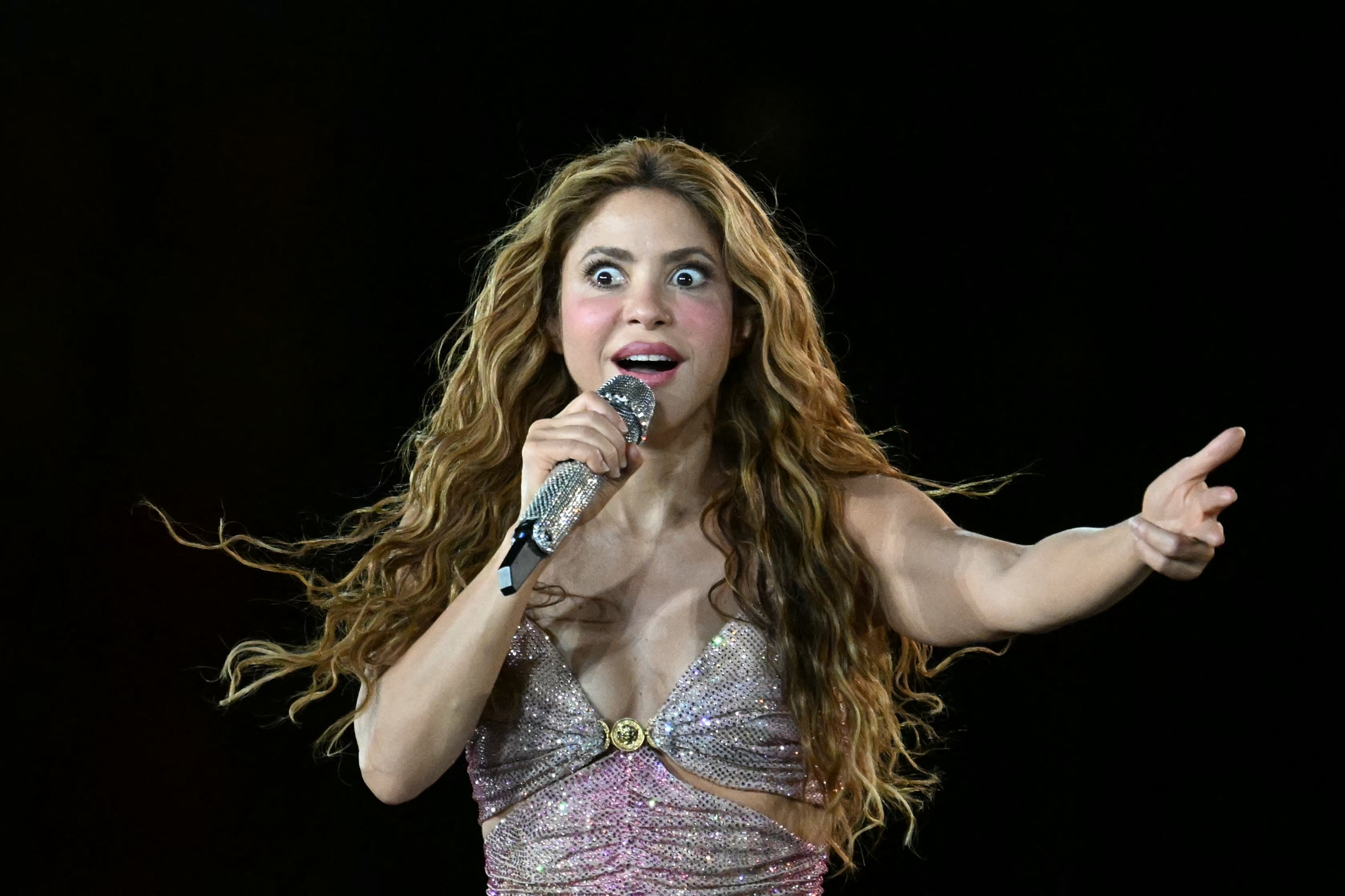 Singer with wavy hair energetically performing on stage, holding a microphone and wearing a sparkly dress with a deep neckline