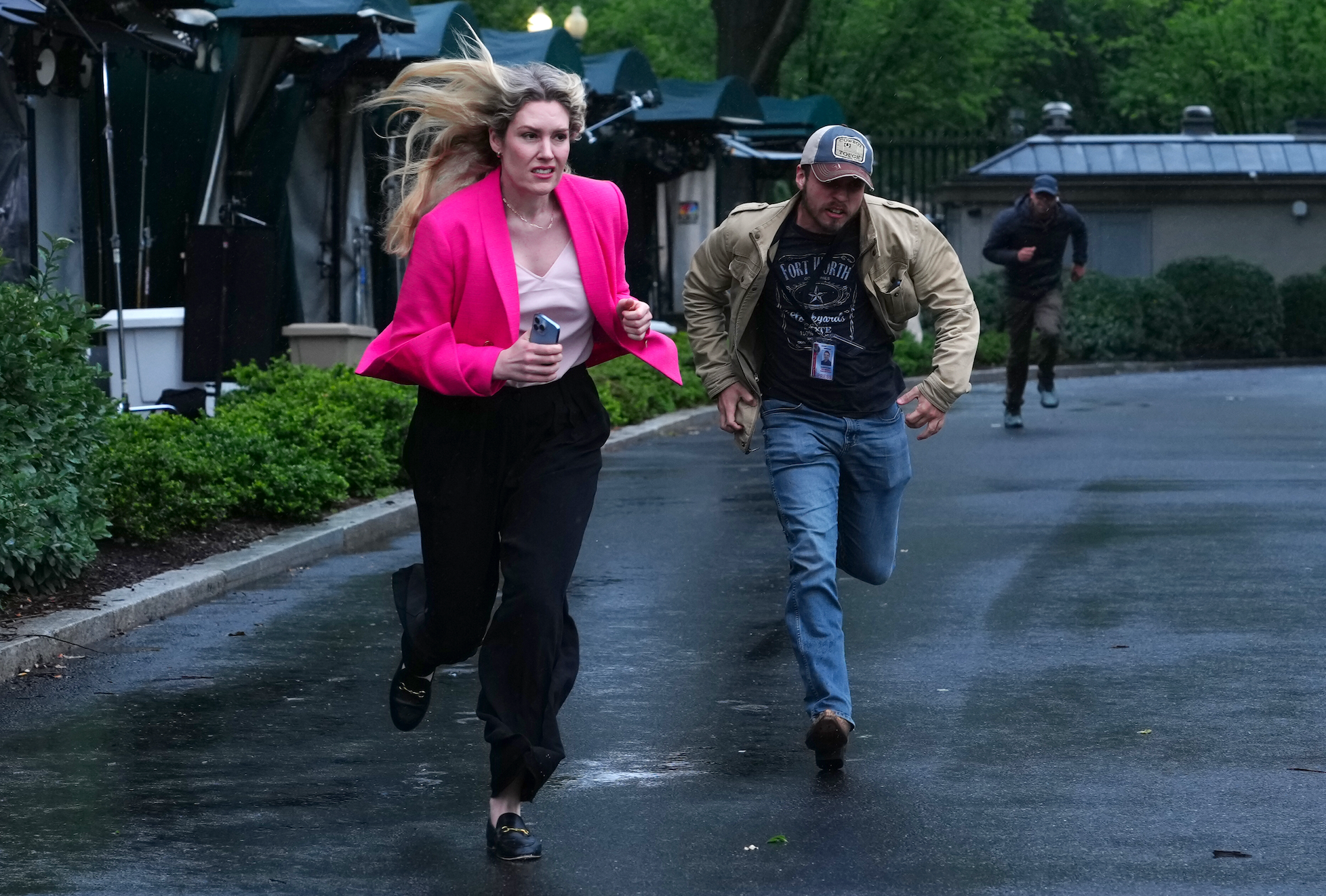 A woman in a stylish blazer runs energetically, holding a cup, pursued by a man in a casual jacket and cap