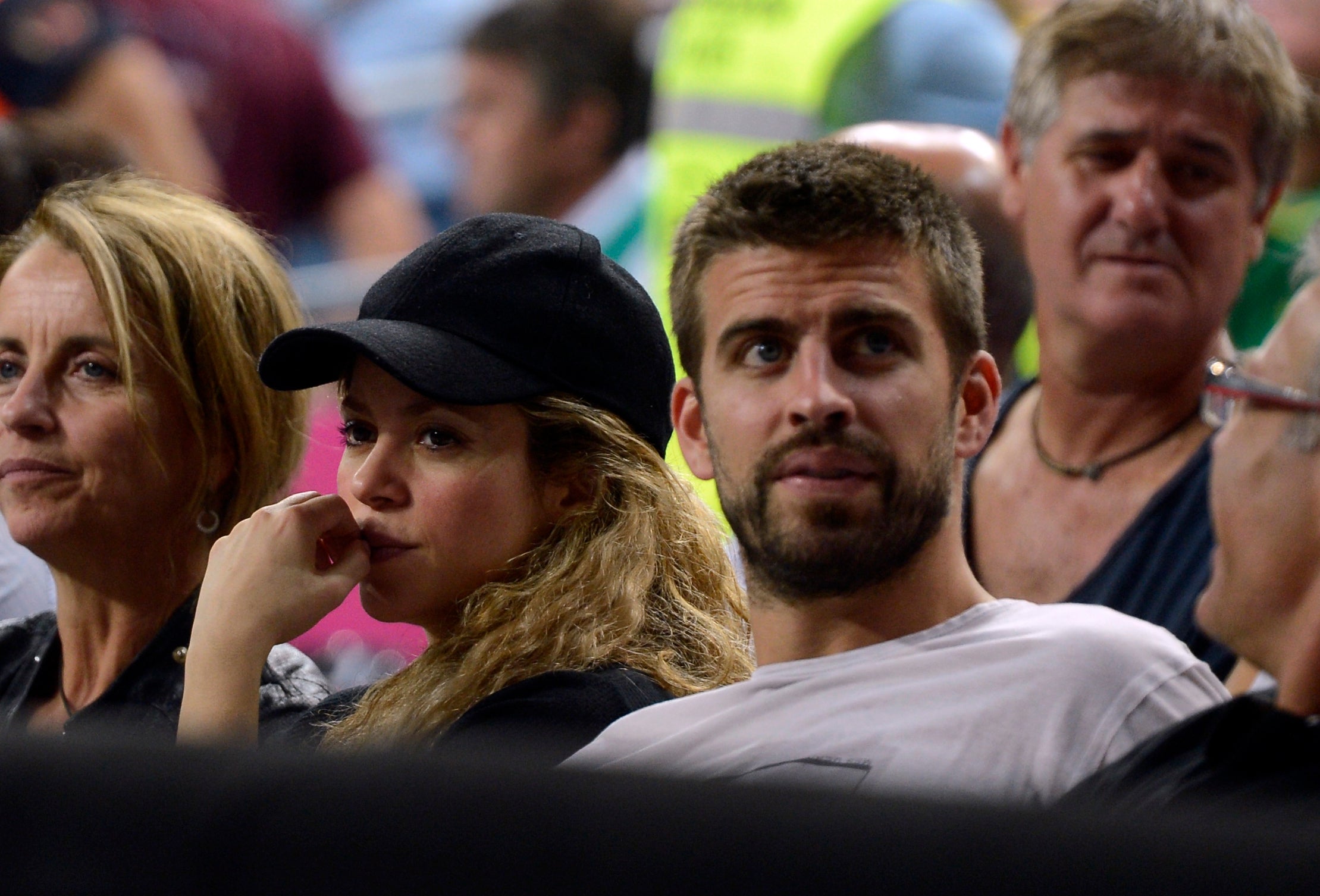 Two people seated at an event, looking towards the action. The woman wears a cap and the man has a shirt. Audience members are in the background