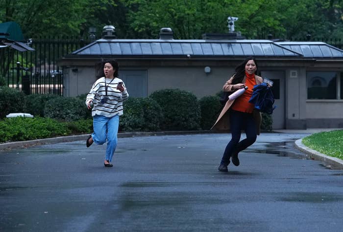 Two people running outdoors on a wet path, appearing hurried or in pursuit of something, with a building and greenery in the background